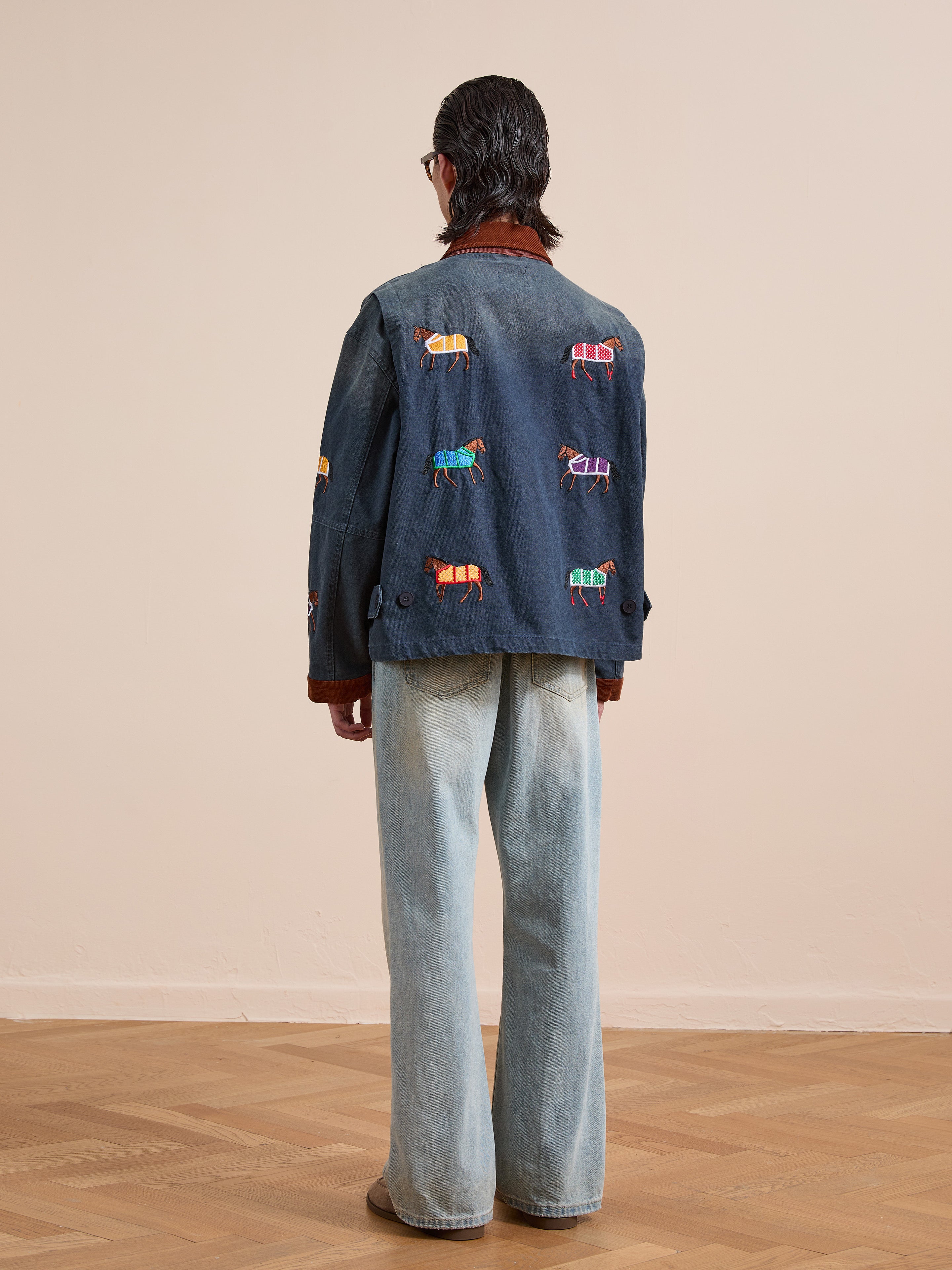 A person with long hair stands facing away, wearing light blue wide-leg jeans and the Found Lariat Horse Embroidered Work Jacket, featuring colorful embroidery, on a wooden floor with a plain beige wall background.