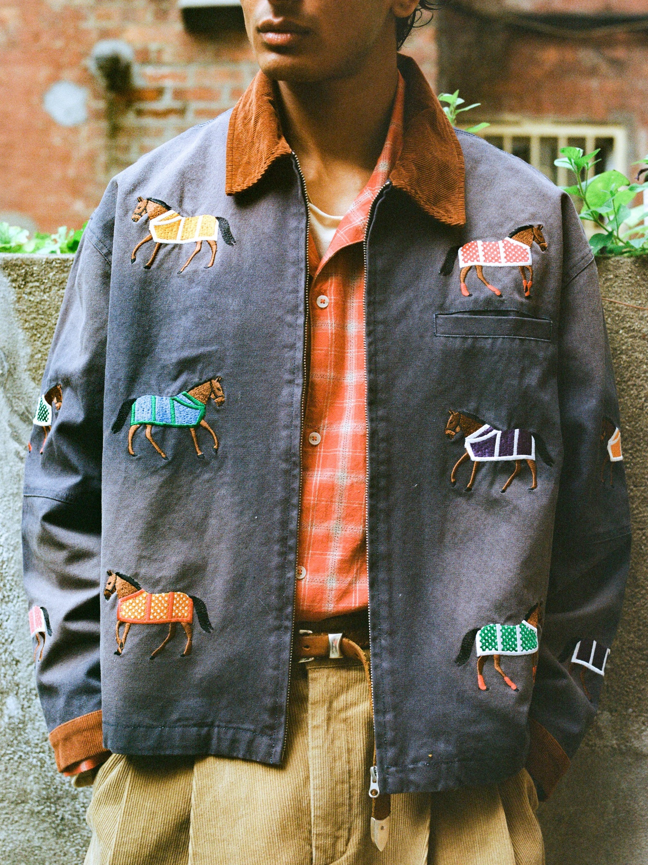 A person stands outdoors in front of a brick wall, wearing Found’s Lariat Horse Embroidered Work Jacket in grey, styled with a plaid shirt and beige corduroy pants.
