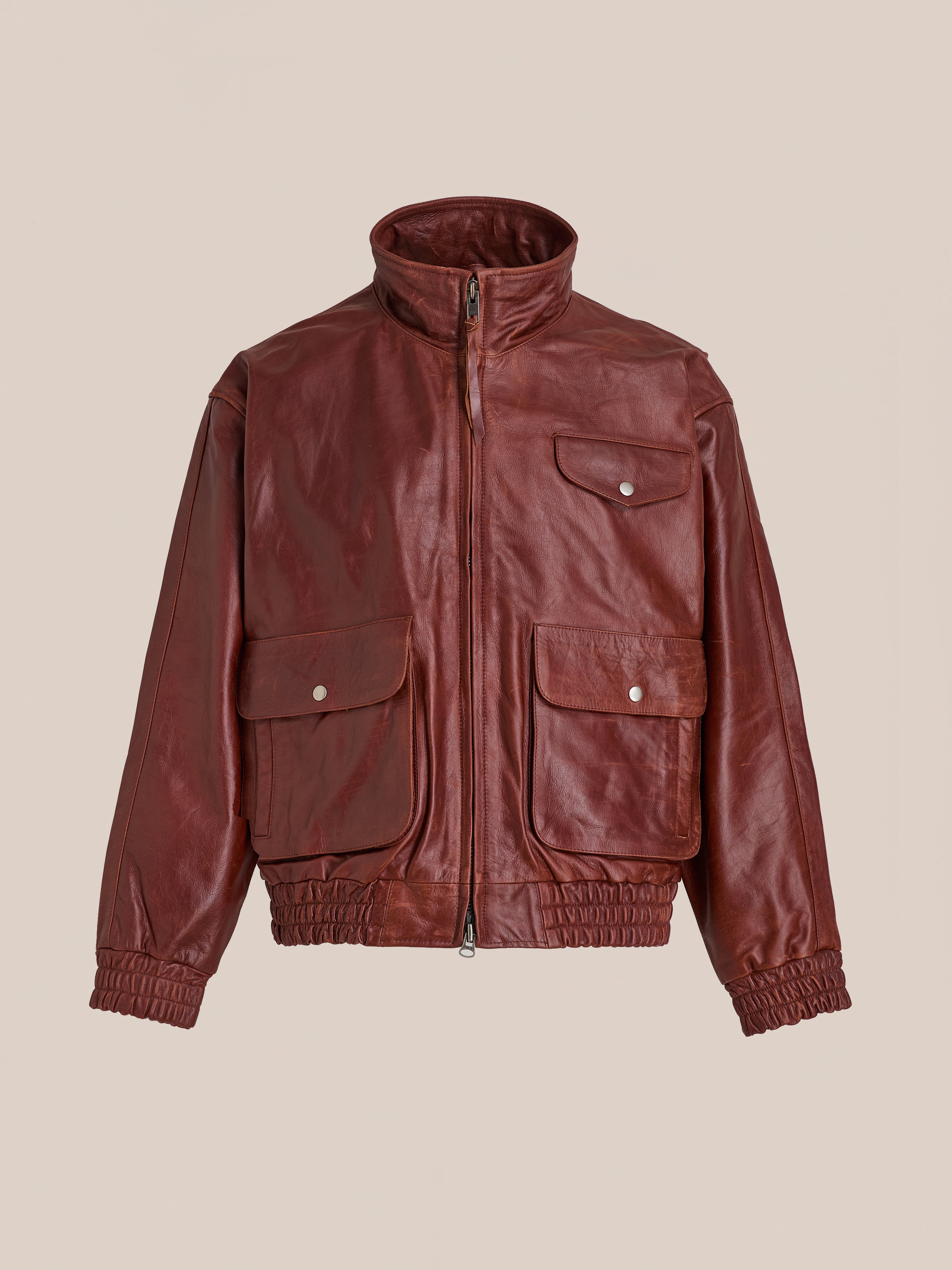 The Redwood Leather Bomber Jacket by Found features brown genuine leather, a front zipper, three snap-button flap pockets, a high collar, and elastic cuffs and waistband. This unisex jacket is shown on a plain light background.