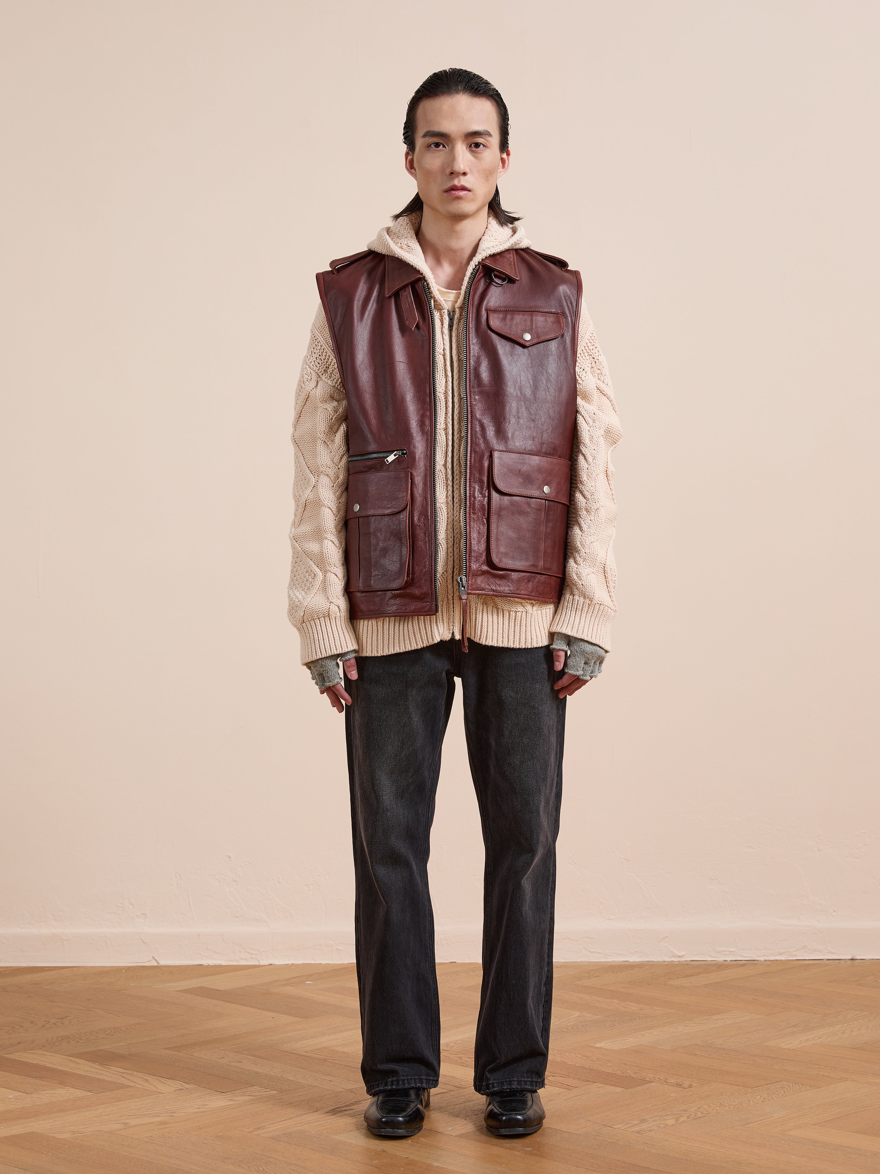Indoors against a light wall and wooden floor, someone wears the Found Leather Pocket Vest with utility pockets over a beige knit sweater, dark jeans, fingerless gloves, and black shoes.