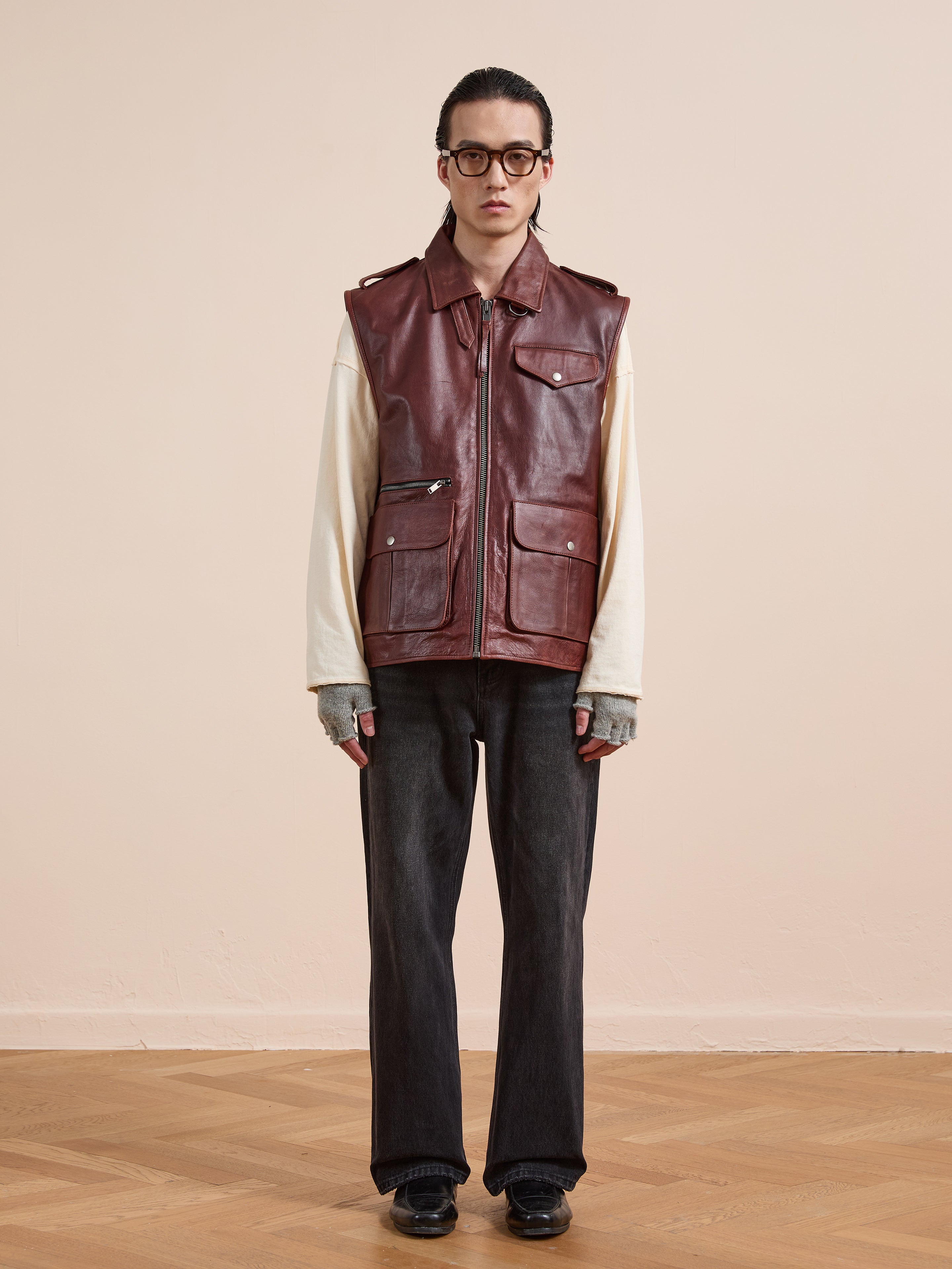 Indoors, a person models the Found Leather Pocket Vest in maroon with utility pockets, styled over a cream long-sleeve shirt, dark jeans, gray fingerless gloves, round glasses, and black shoes.