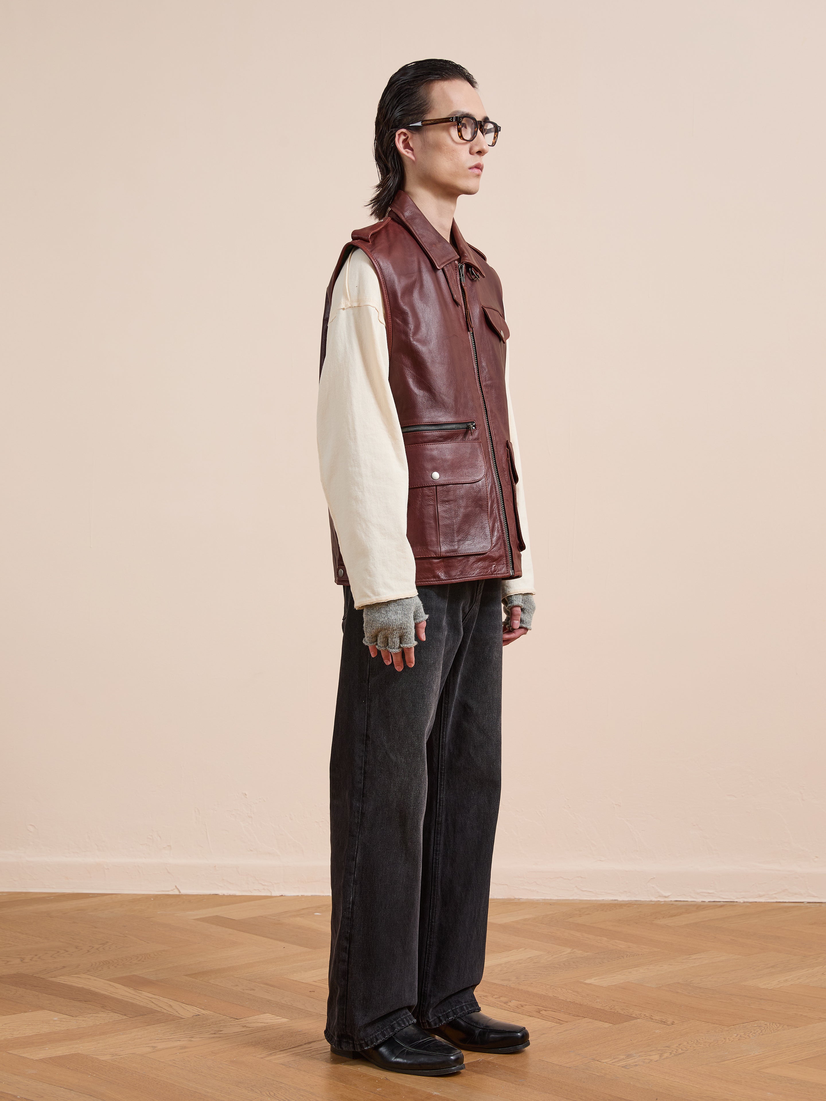 A person with long hair, glasses, and gloves wears the Found Leather Pocket Vest in maroon over a cream shirt with wide black jeans and black shoes, standing on a wooden floor against a beige wall.