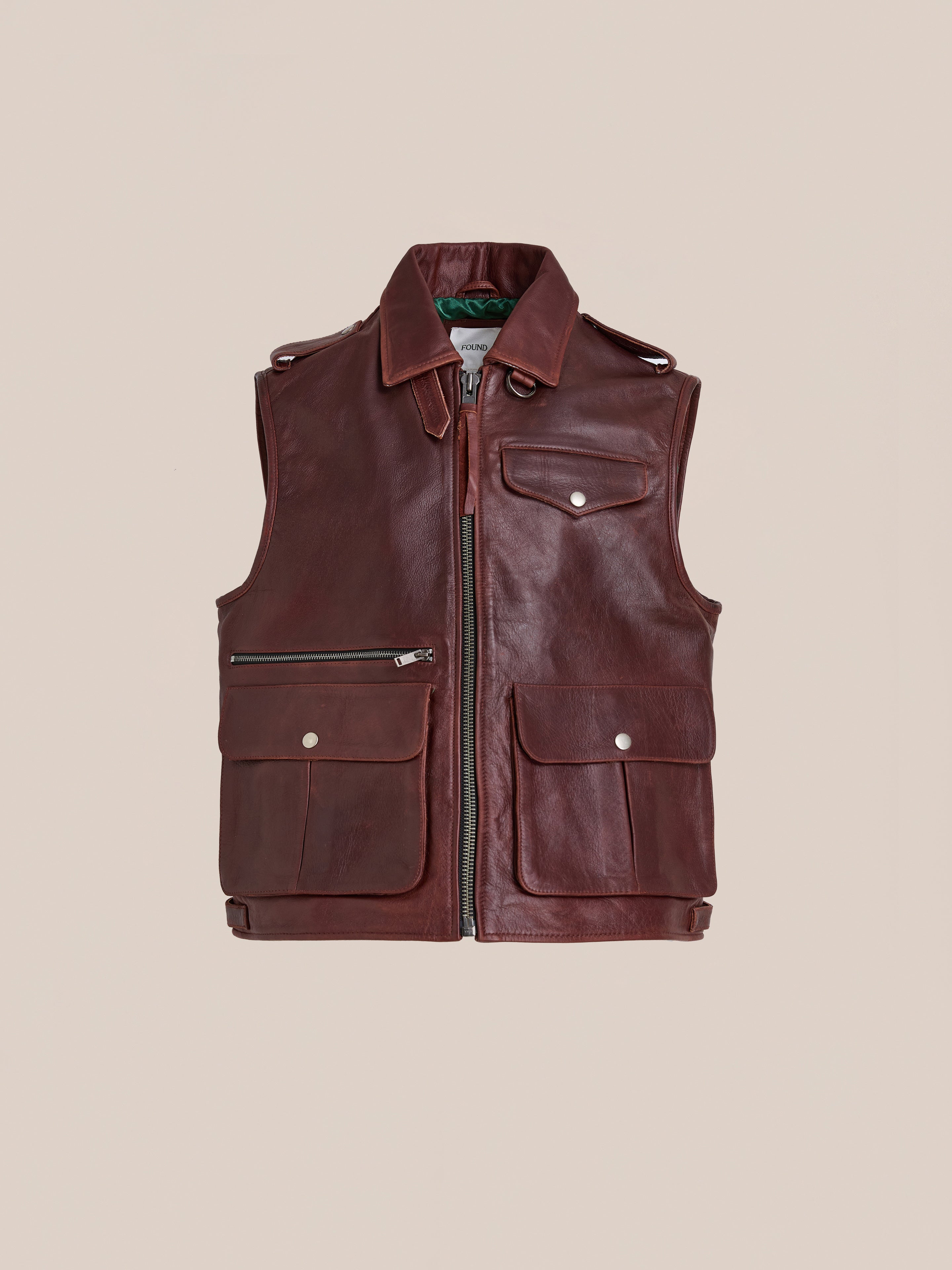 The Found Leather Pocket Vest, made from distressed brown leather, features a front zipper, utility pockets with flaps and zippers, and epaulet details. Shown against a plain beige background.