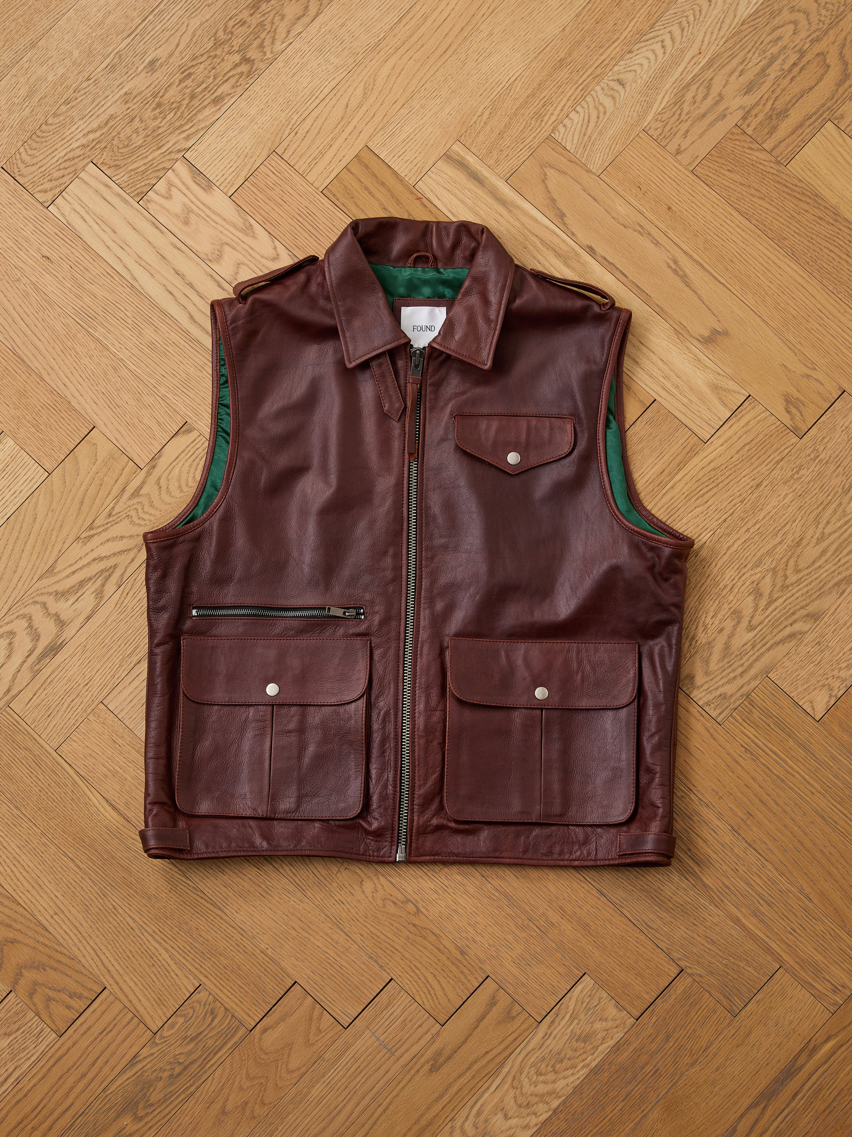 The Found Leather Pocket Vest in distressed brown leather features utility pockets, a front zipper, zippered chest pocket, and green inner lining, shown laid flat on a wooden parquet floor.