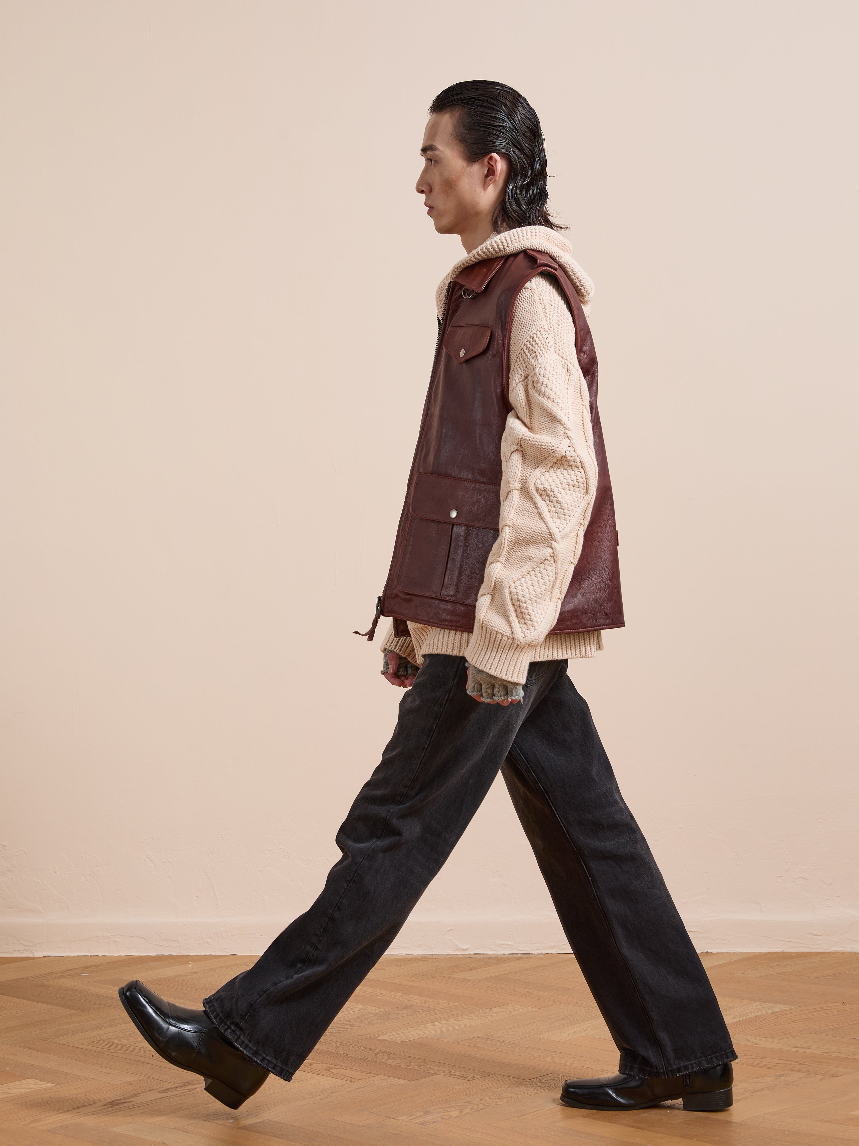 A person wearing Found’s Leather Pocket Vest with utility pockets over a cream knit sweater, paired with dark wide-leg pants and black shoes, is photographed in profile against a beige wall and wooden floor.
