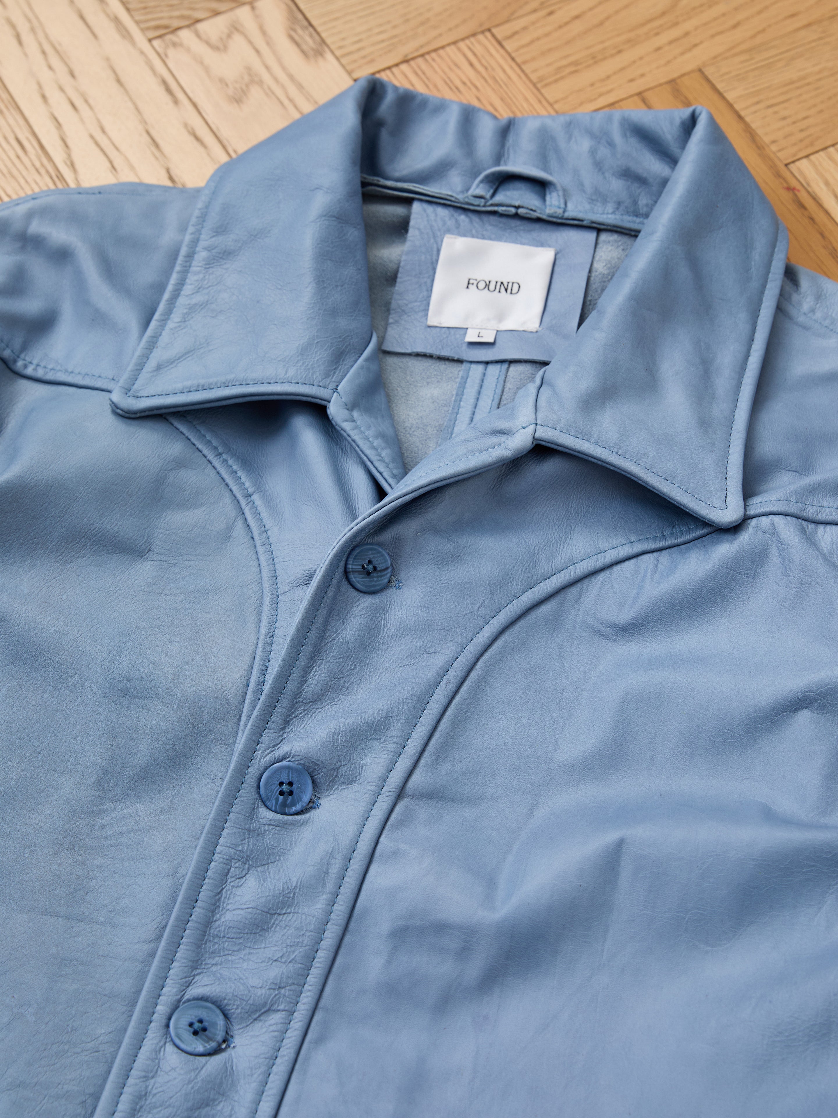 A light blue unisex Leather Field Jacket by FOUND with a collar is laid flat on a wooden floor. A white label inside reads "FOUND.