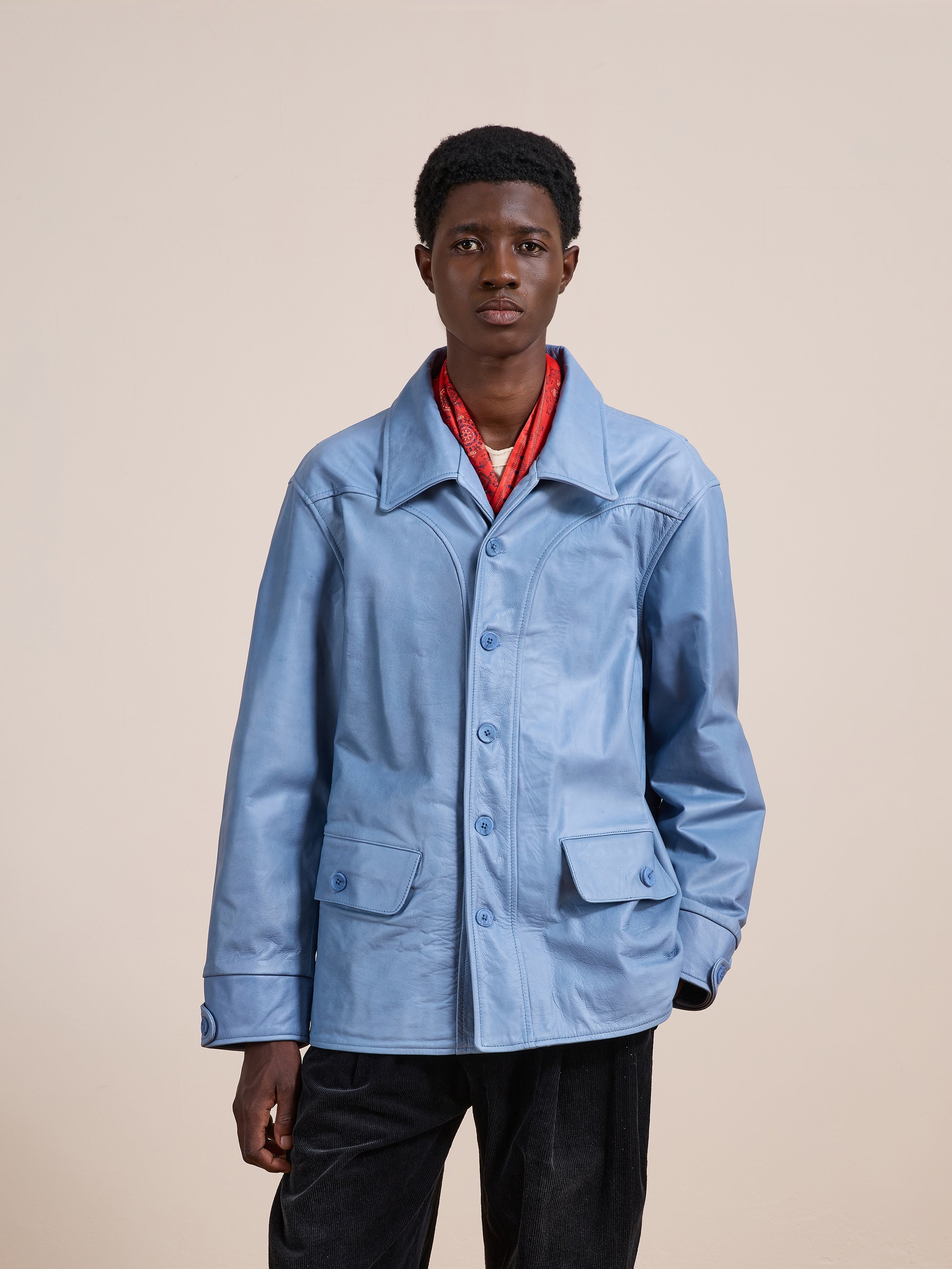 A person stands against a plain backdrop wearing the FOUND Leather Field Jacket, which features a relaxed unisex fit in light blue, paired with a red scarf and dark pants. They face the camera with a neutral expression.