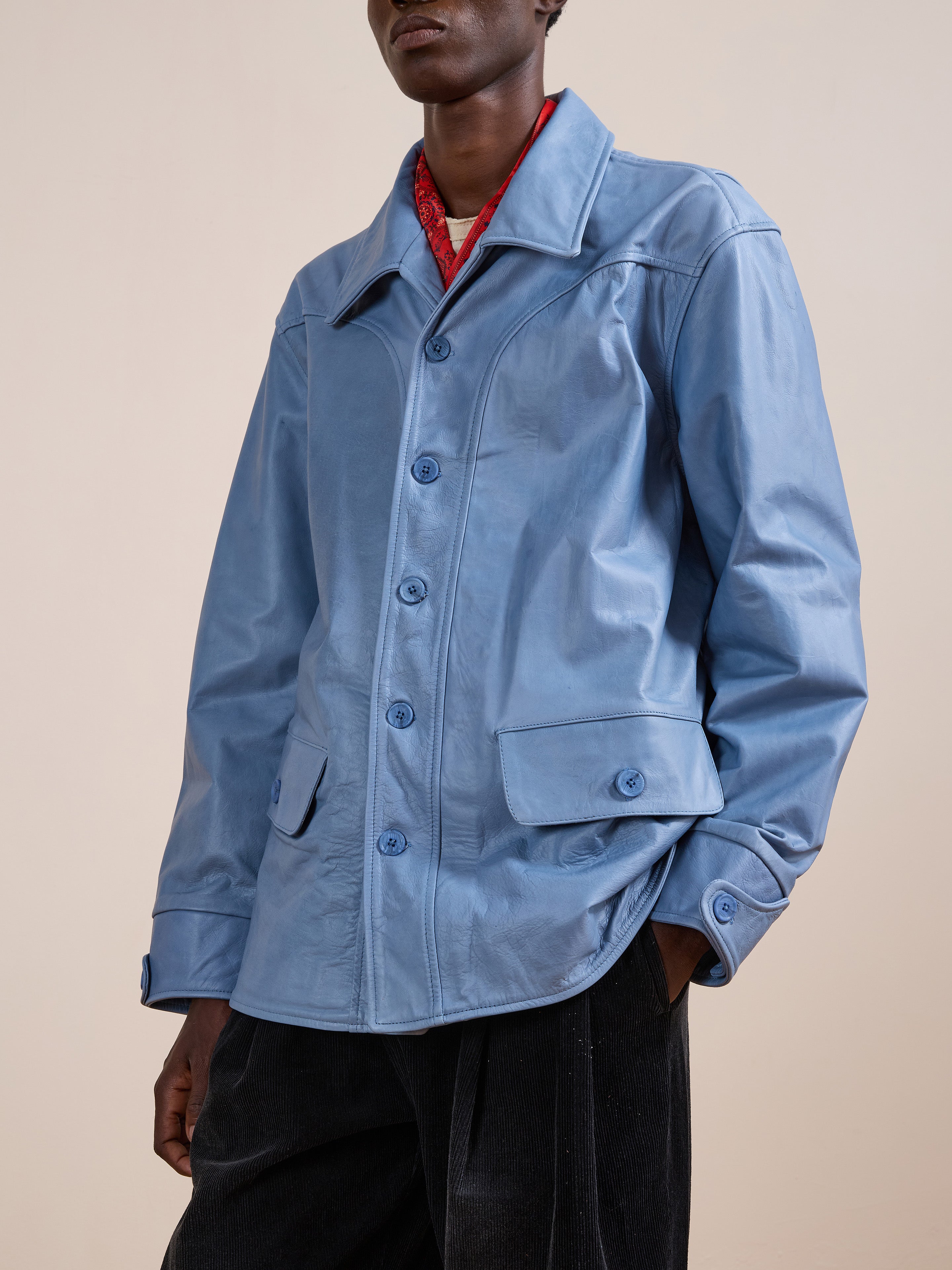 Model wears FOUND's Leather Field Jacket in light blue, relaxed fit, over a red shirt with hands in pockets, standing against a plain beige background.