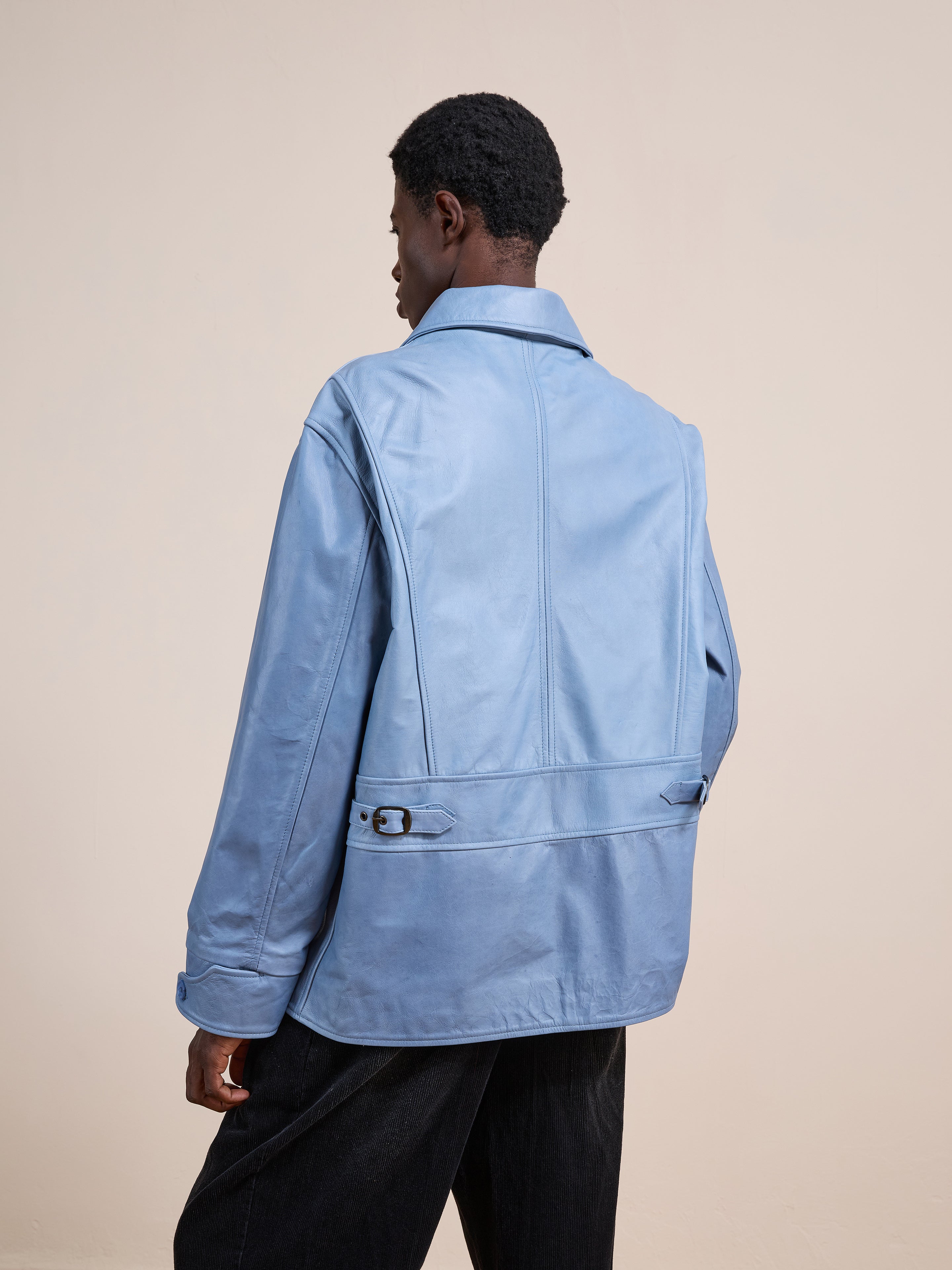 A person wearing a relaxed fit FOUND Leather Field Jacket in light blue and black pants stands with their back to the camera against a plain beige background.