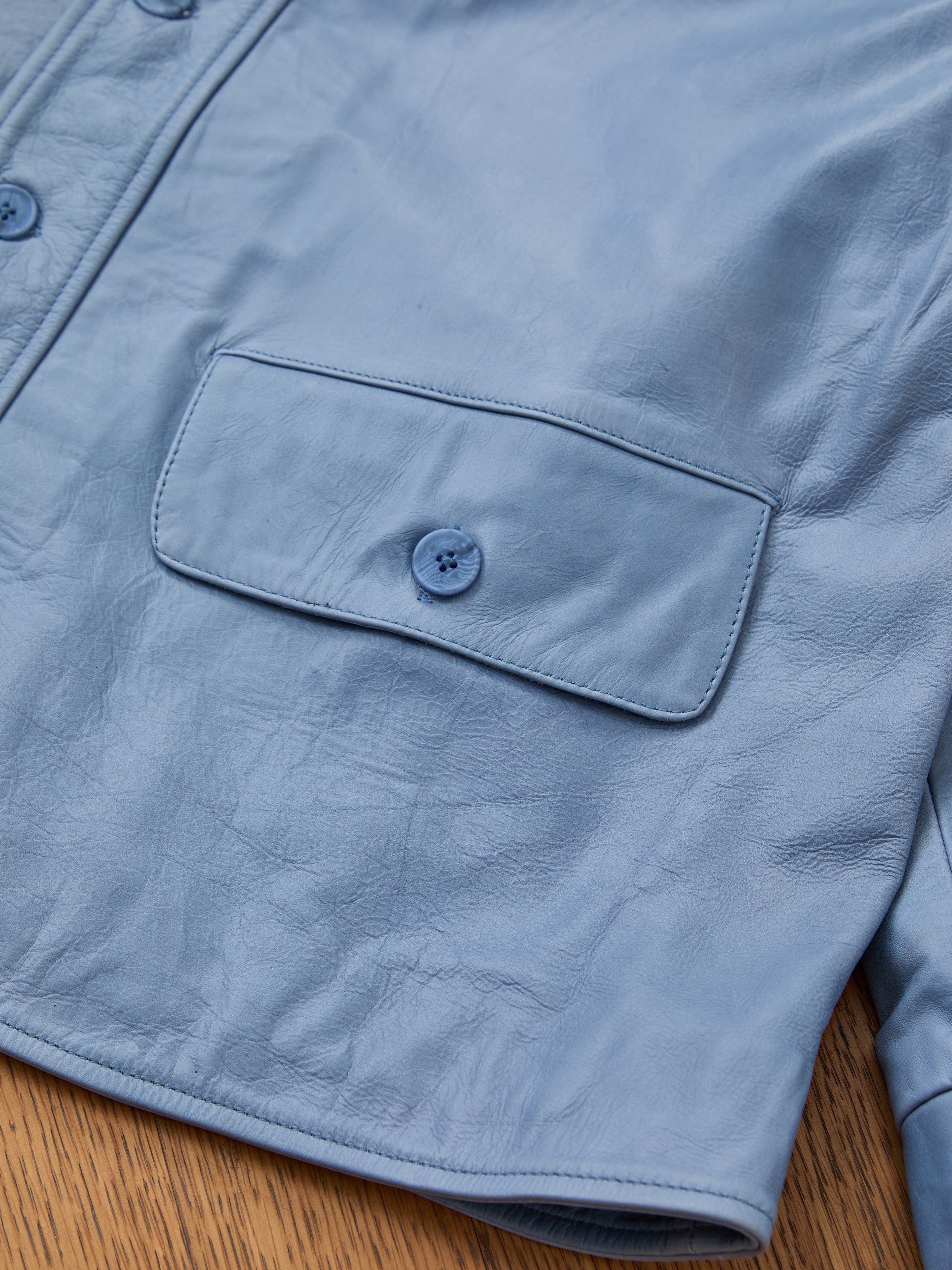 Close-up of the FOUND Leather Field Jacket, a unisex light blue piece with textured leather, a buttoned flap pocket, and partial button front detail, displayed on a wooden surface.