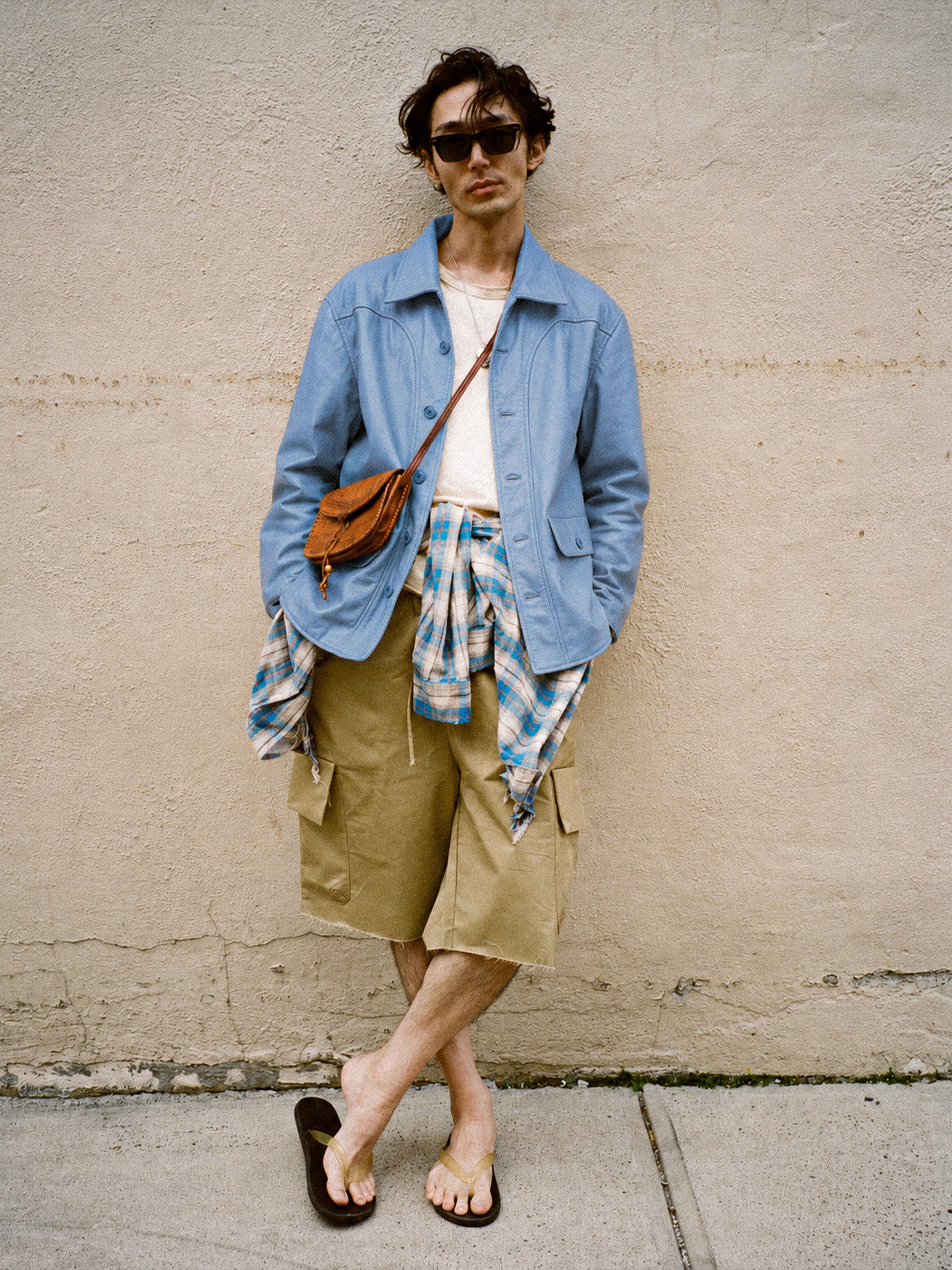 A person stands against a beige wall wearing sunglasses, the FOUND Leather Field Jacket in a relaxed unisex fit, a plaid shirt tied at the waist, tan cargo shorts, flip-flops, and a small brown crossbody bag.