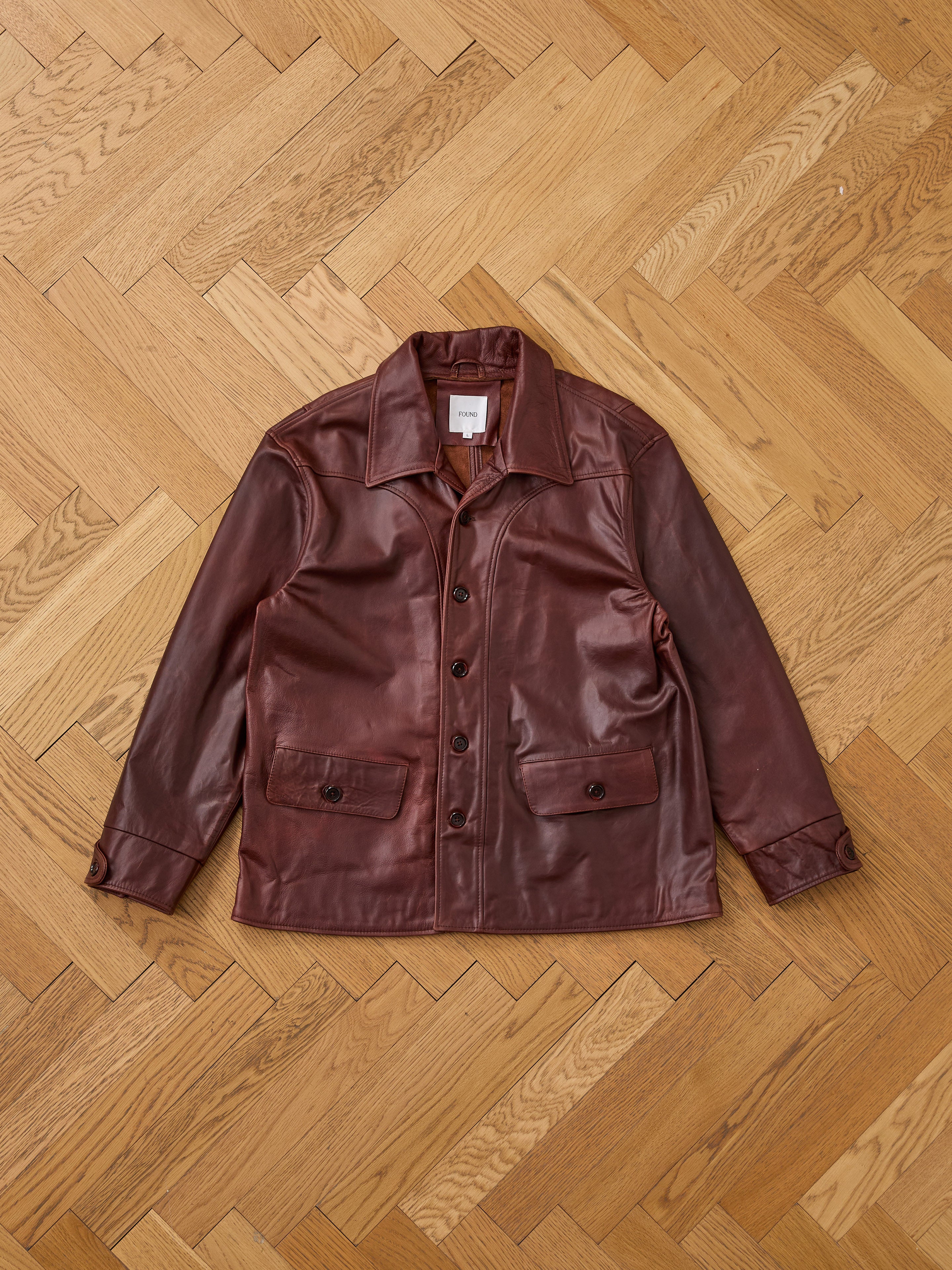The FOUND Leather Field Jacket is a relaxed fit, unisex brown leather button-up with a collar and two front pockets, shown laid flat on a wooden herringbone floor.