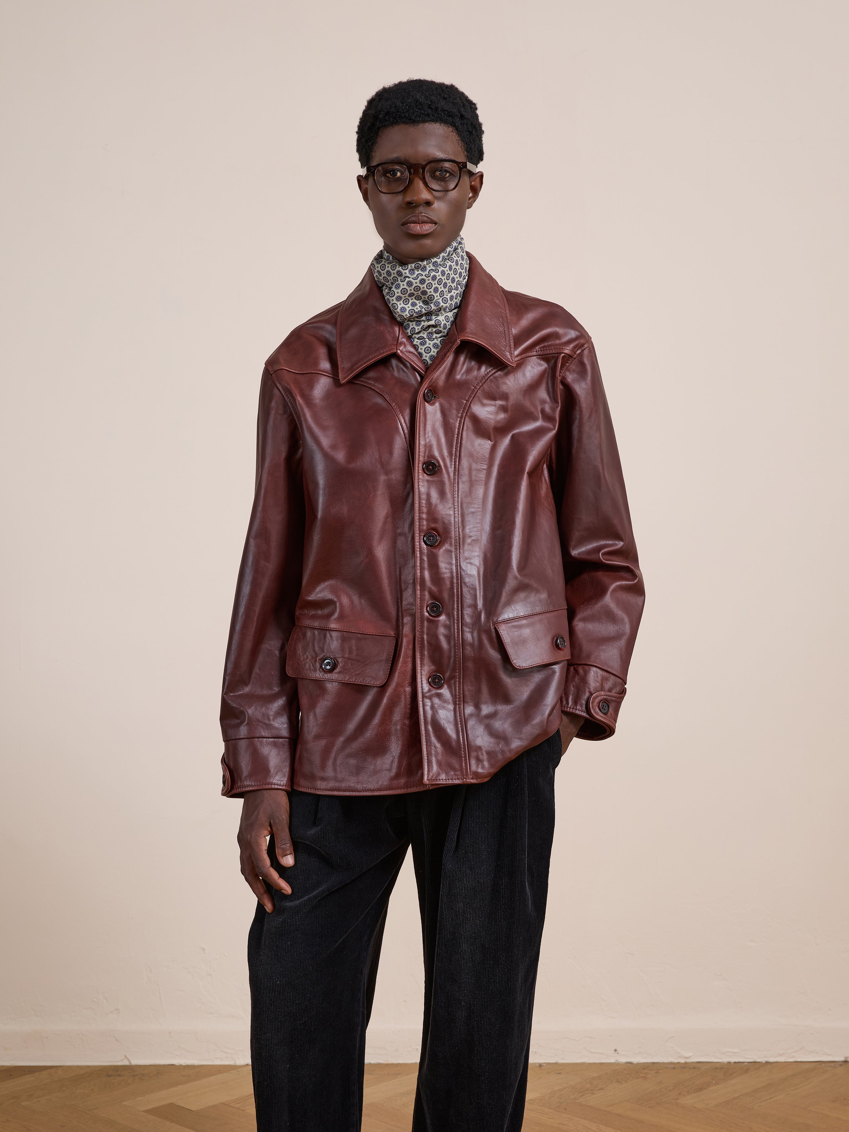 A person wearing glasses, a patterned scarf, black pants, and the FOUND Leather Field Jacket stands against a plain beige wall.