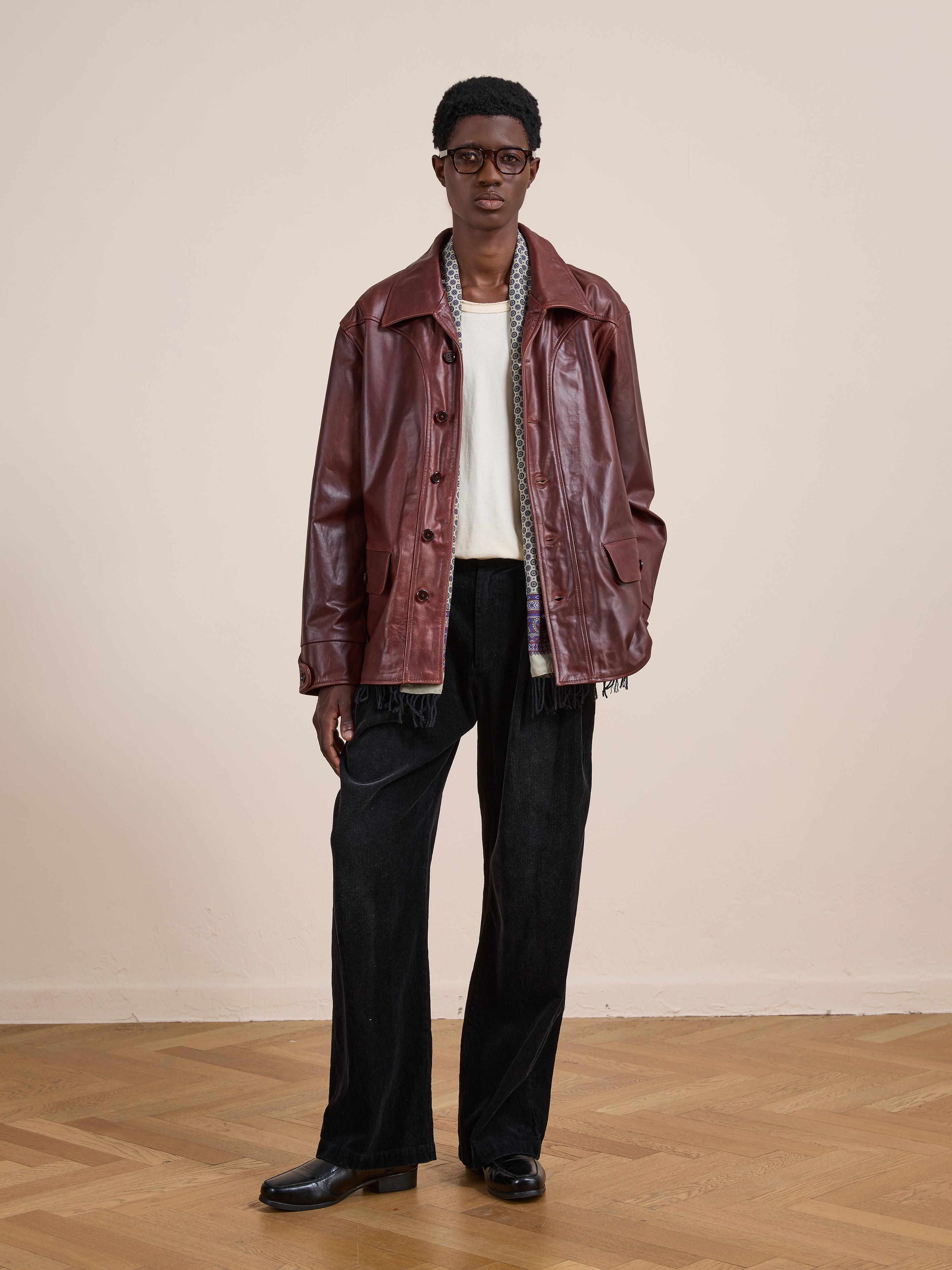 A person stands indoors wearing the FOUND Leather Field Jacket in brown, paired with a cream shirt, black pants, black shoes, and glasses on a parquet floor against a plain beige wall.