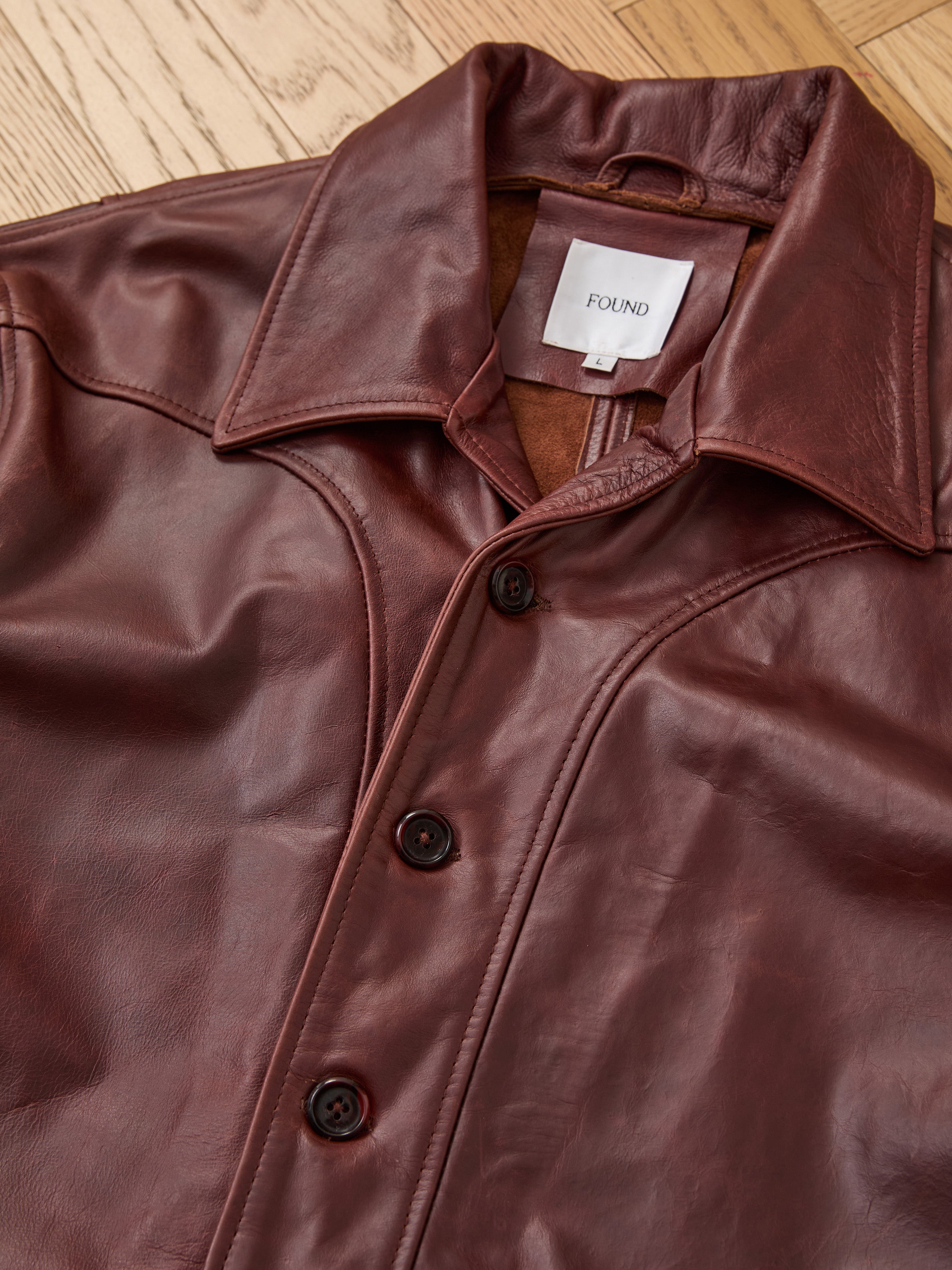 The FOUND Leather Field Jacket is a brown unisex piece featuring a relaxed fit, large collar, and three dark buttons, shown laid flat on a wooden floor with the "FOUND" label inside.