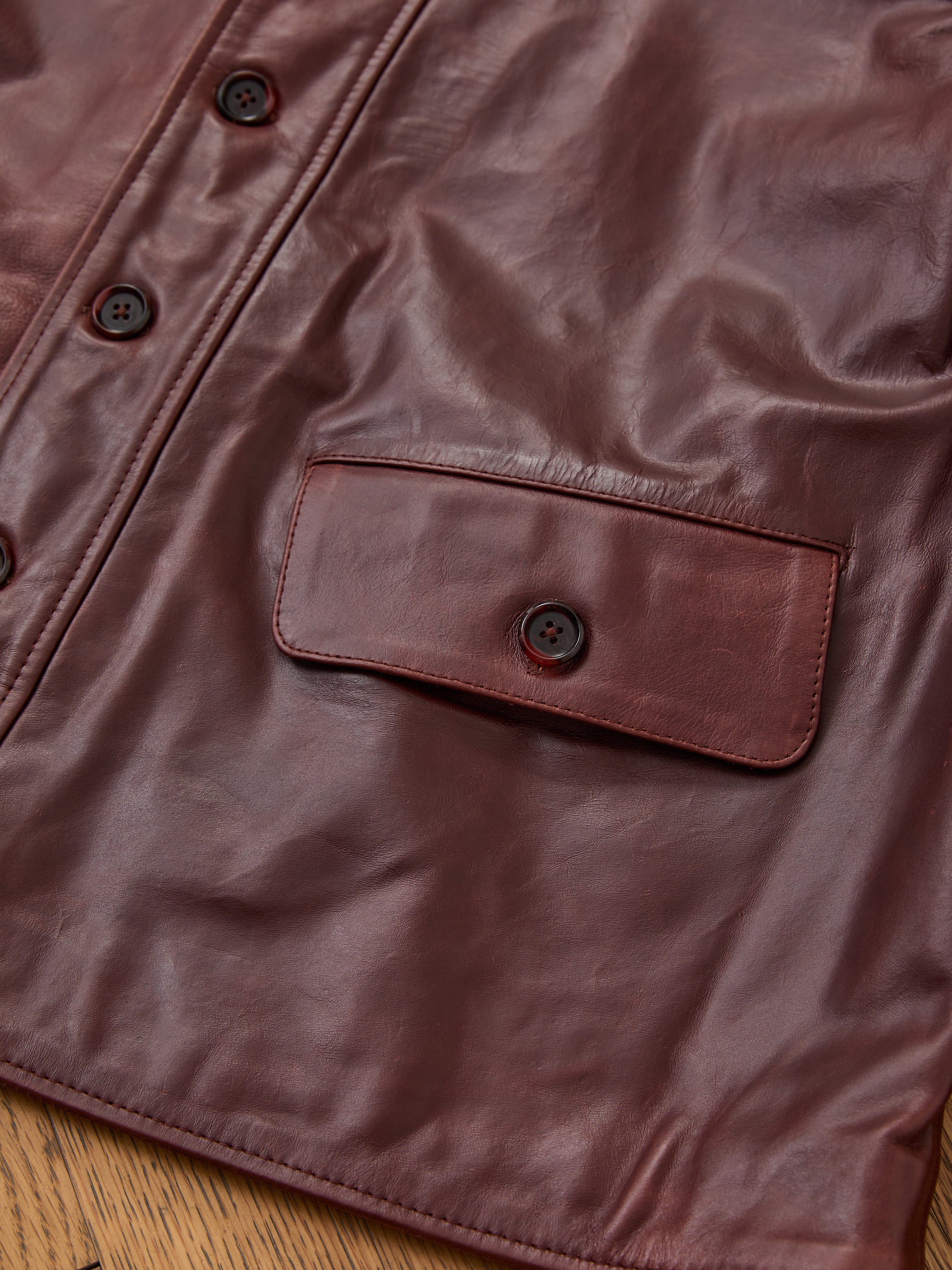 Close-up of the FOUND Leather Field Jacket, a unisex brown design with a buttoned flap pocket and partial buttoned front placket, displayed on a wooden surface.