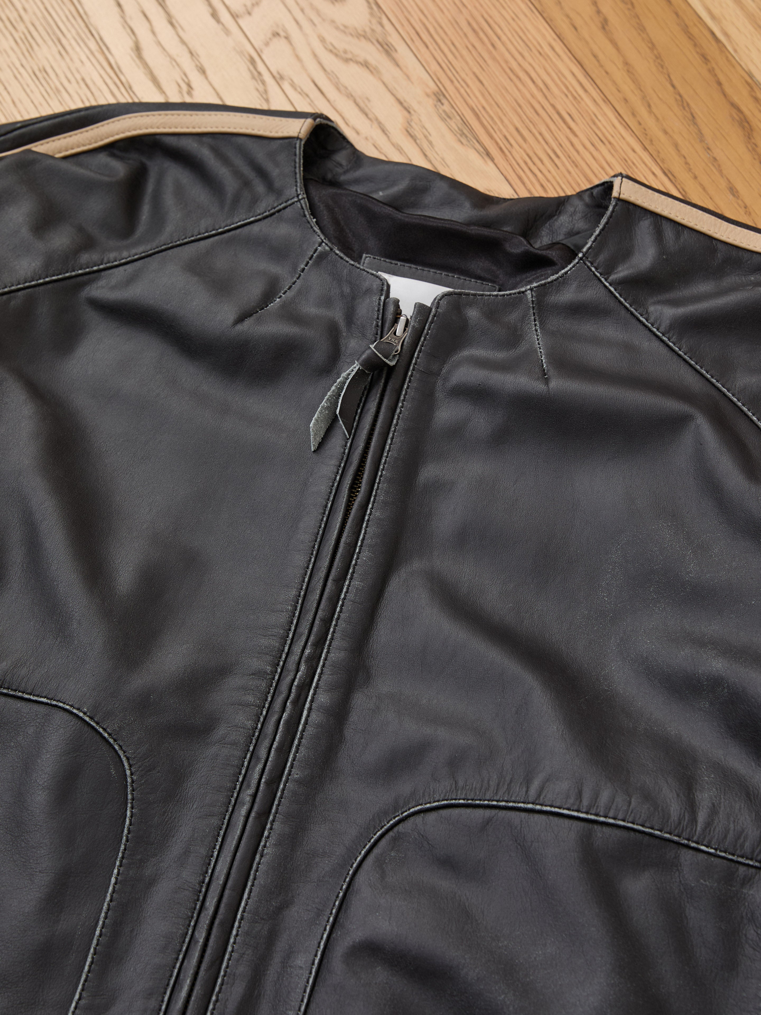 The FOUND Leather Track Jacket is laid flat on a wooden floor, featuring a front zipper, visible stitched details, and a smooth leather texture.