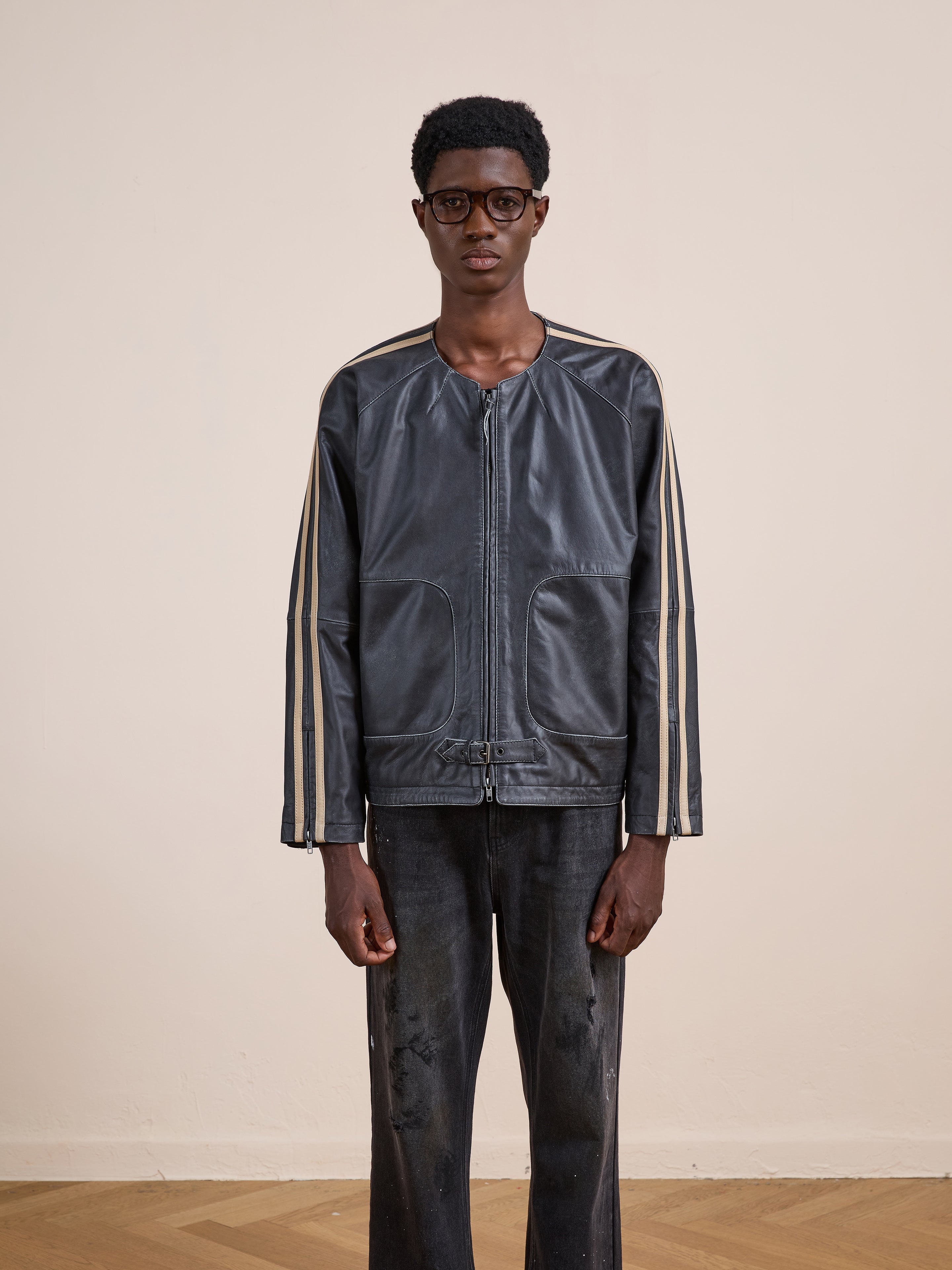 Indoors against a plain beige background, a person wears glasses, distressed black pants, and the FOUND Leather Track Jacket—unisex style with beige sleeve stripes.