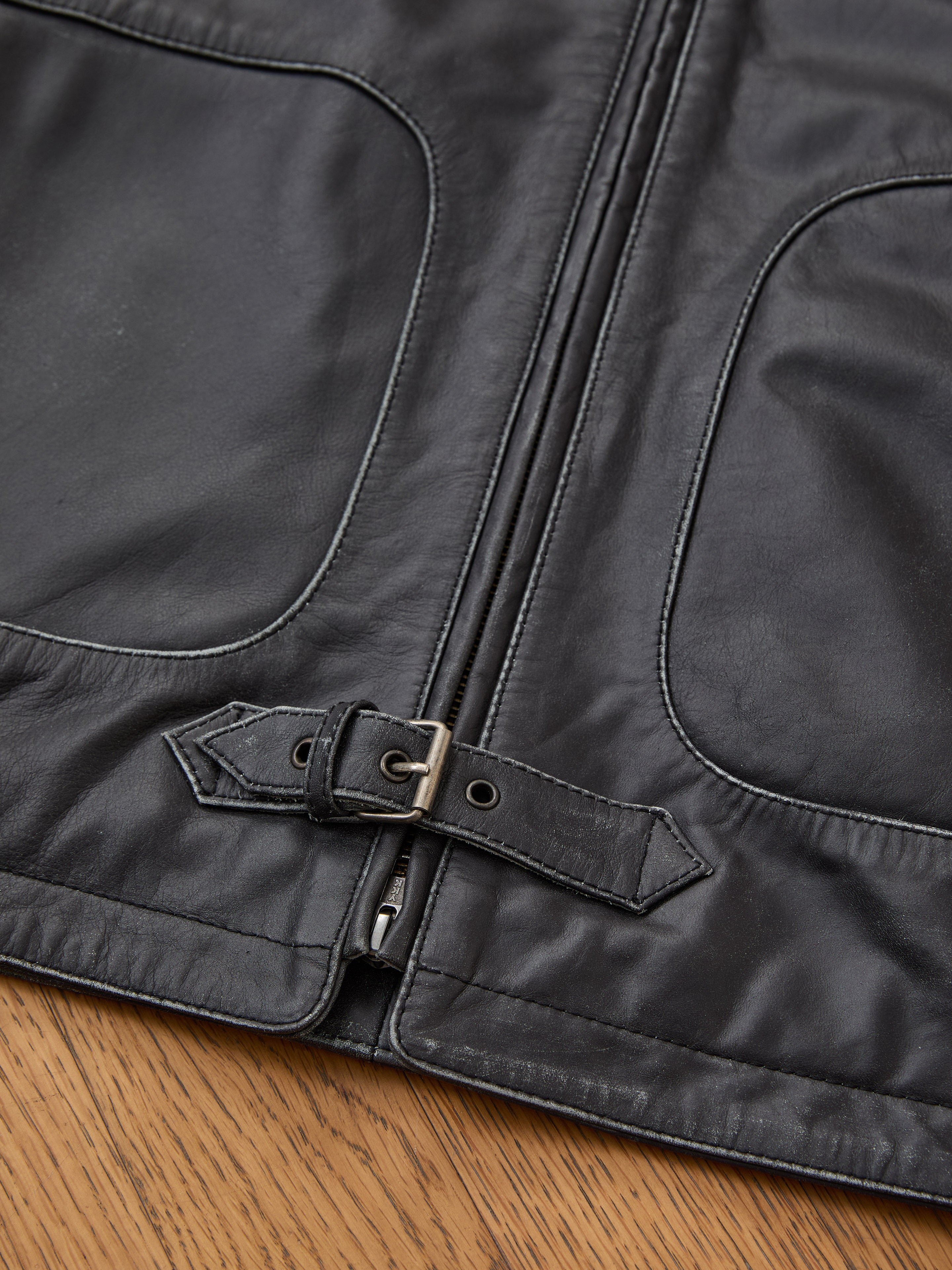 Close-up of the FOUND Leather Track Jacket in black, shown on a wooden surface; features two pockets, a zipper, and a small belted strap at the hem.