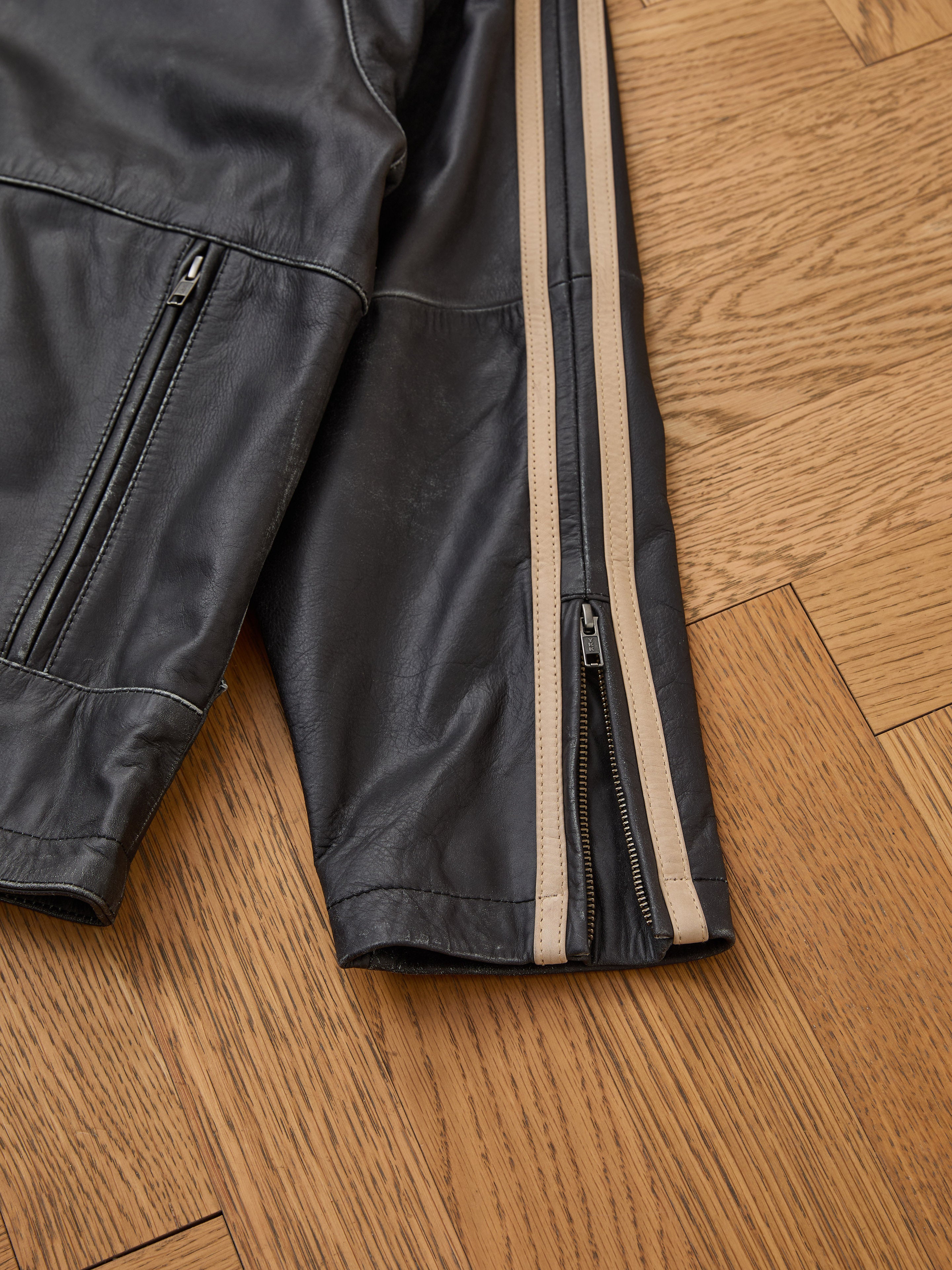 FOUND Leather Track Jacket in black, unisex design, features a beige stripe detail on the sleeve and a zipper front, evoking vintage moto style. Displayed on a wooden floor.