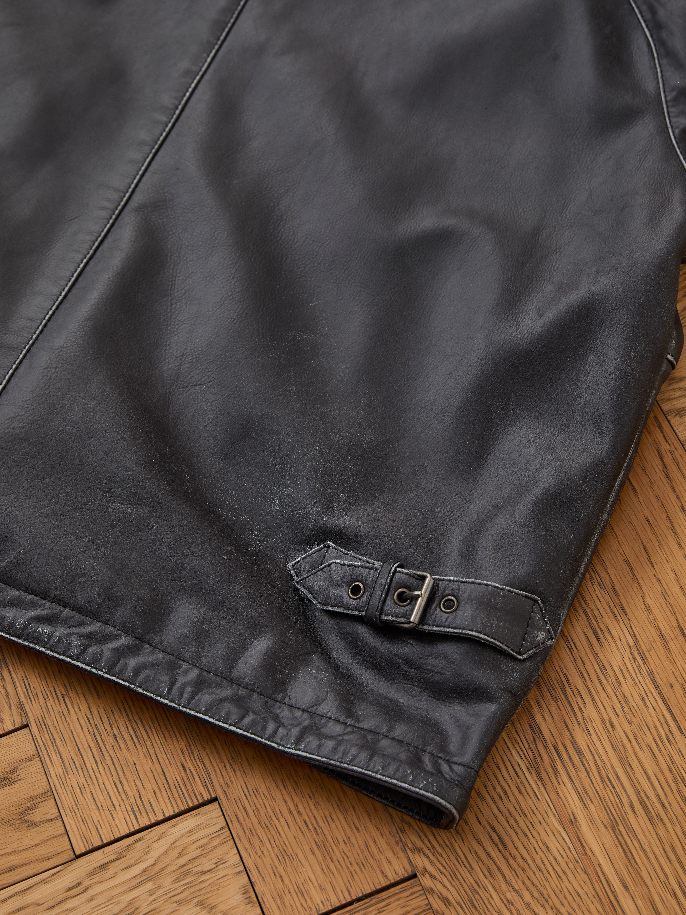 Close-up of the FOUND Leather Track Jacket in black, a unisex design featuring a small belt and buckle detail near the hem, displayed on a wooden floor.