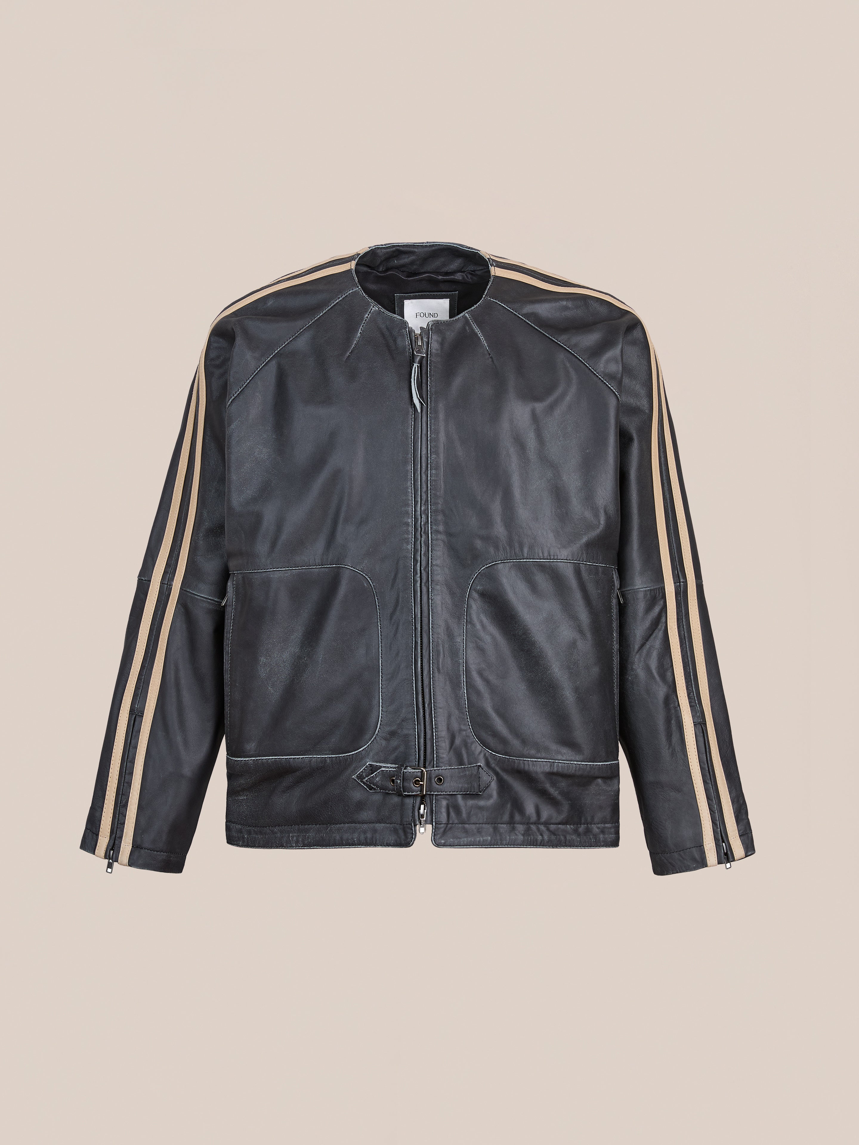 The FOUND Leather Track Jacket is a unisex black leather jacket with tan sleeve stripes, front zipper, two large pockets, and a waist buckle—blending classic style with bold details.