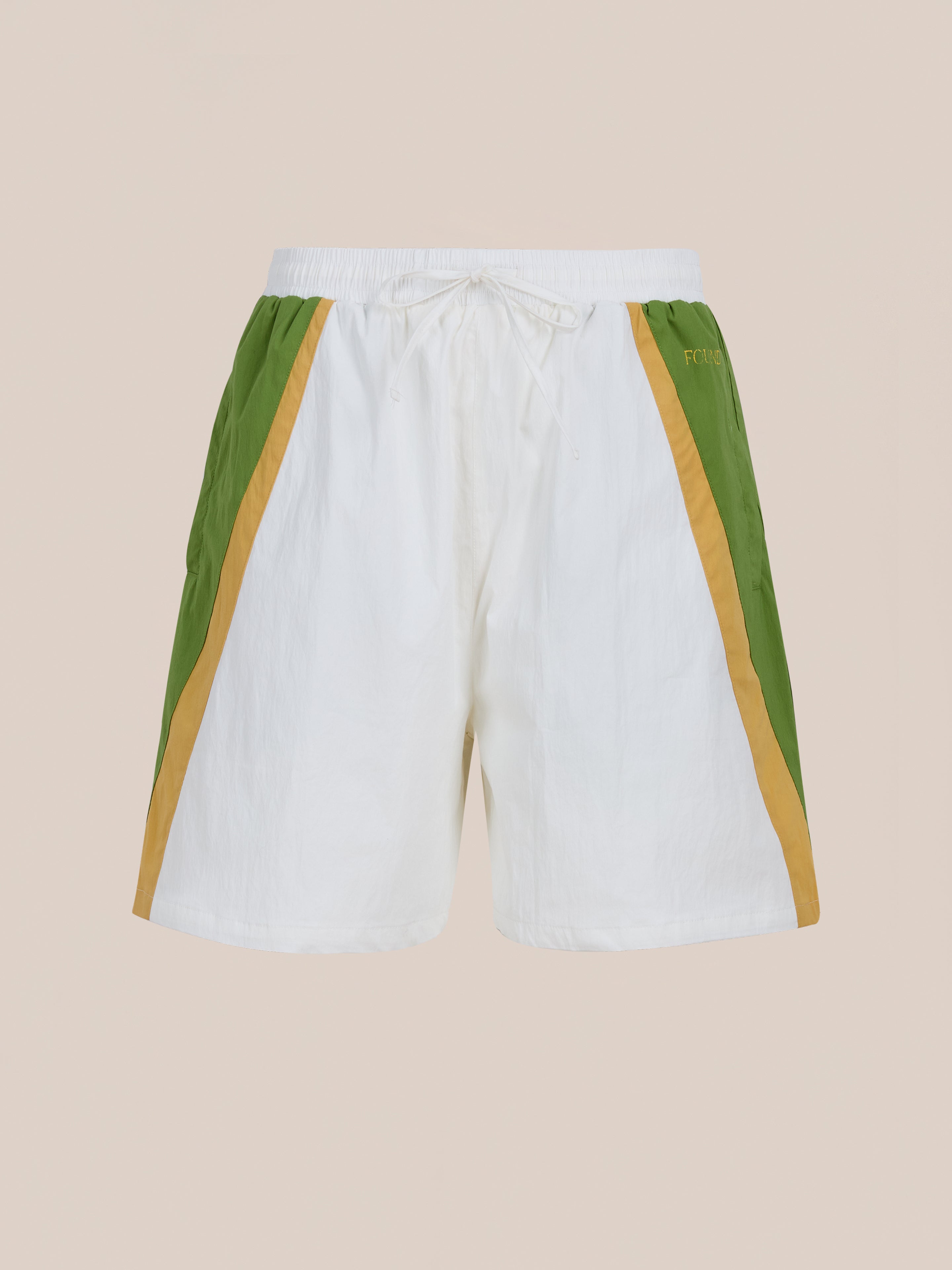 The Legacy Cricket Track Shorts by Found are white unisex athletic shorts with green and tan side panels, an elastic waistband, and a drawstring. Vintage-inspired and perfect for cricket track shorts fans. Shown on a plain beige background.