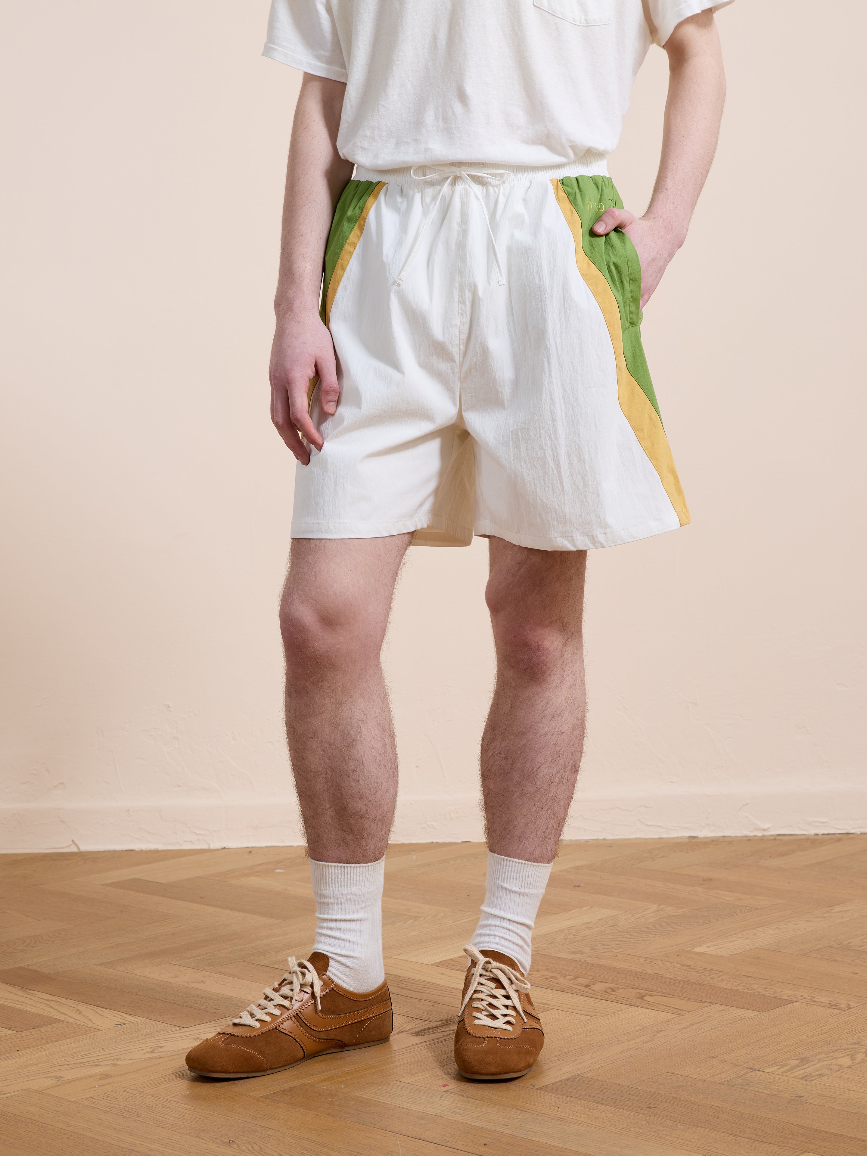 Person wearing Found Legacy Cricket Track Shorts with green and yellow stripes, a white t-shirt, white socks, and brown sneakers, standing on a wooden floor.