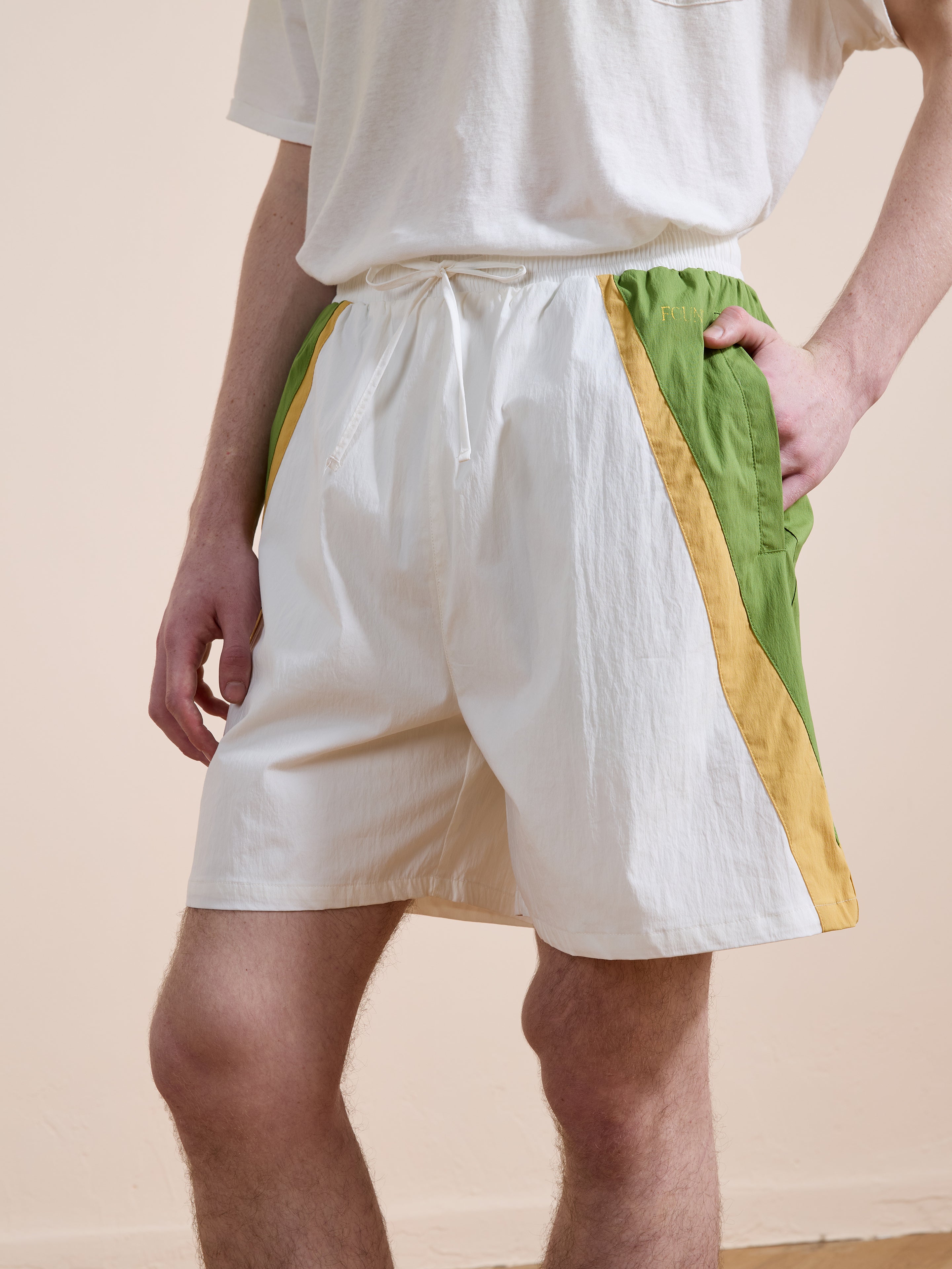 A person wearing a white T-shirt and Found Legacy Cricket Track Shorts with green and yellow side panels, standing with one hand in the shorts pocket.
