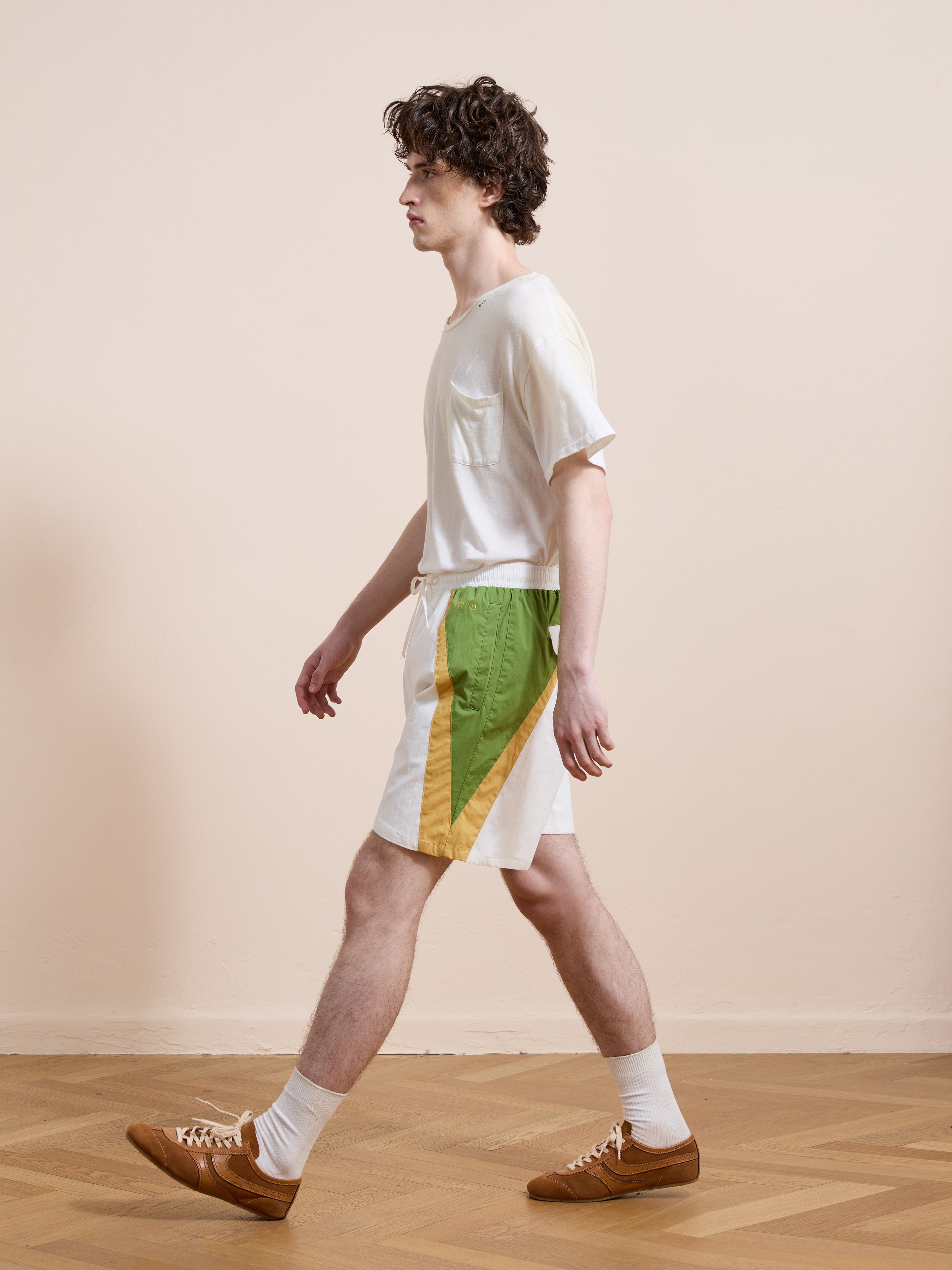 A young man in a white t-shirt, Found Legacy Cricket Track Shorts in white and green, white socks, and brown shoes walks indoors on a parquet floor with a plain beige wall background.