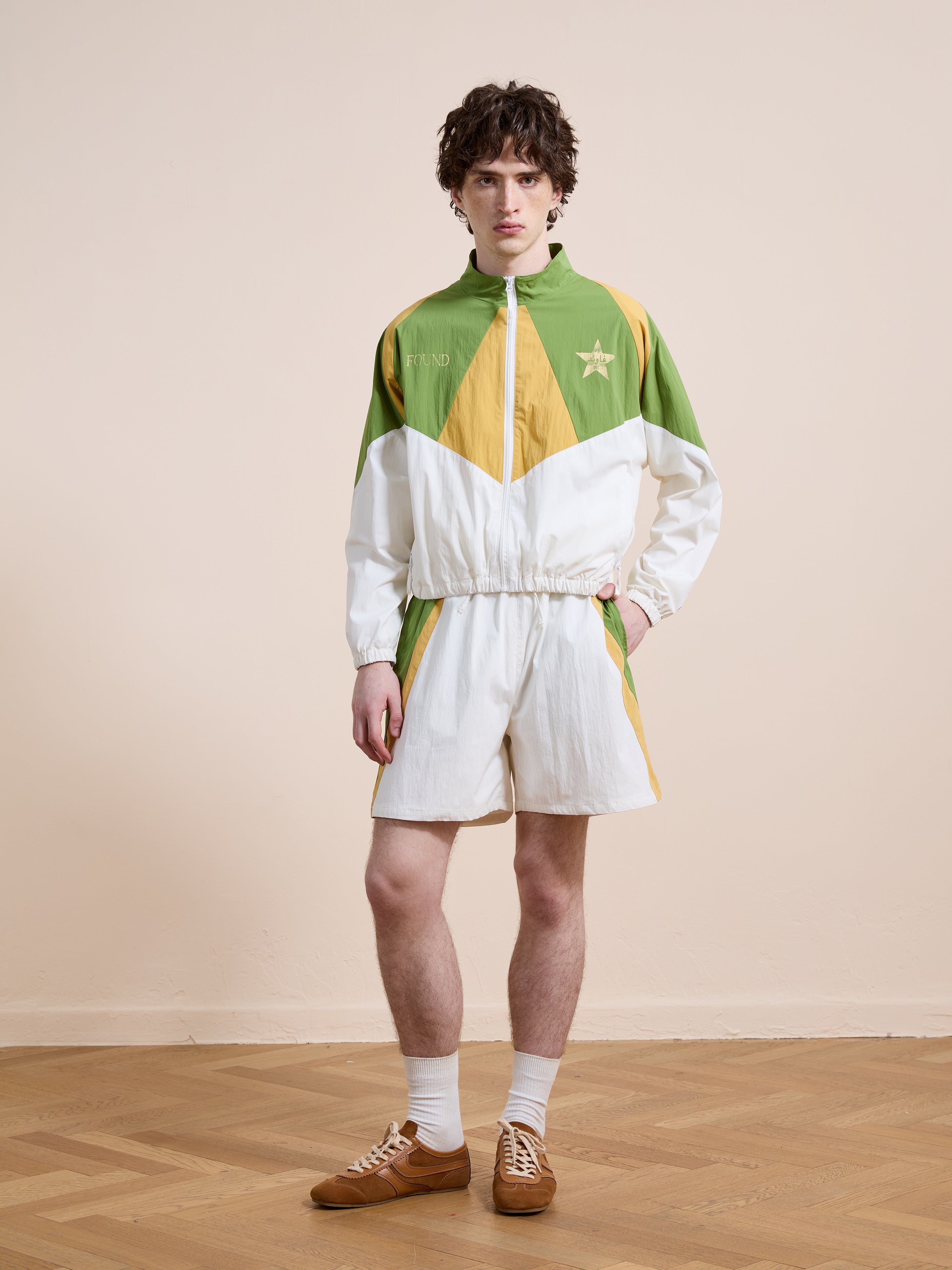 A person stands indoors on a wooden floor wearing Found’s Legacy Cricket Track Shorts, paired with a matching vintage track jacket, white socks, and brown sneakers.