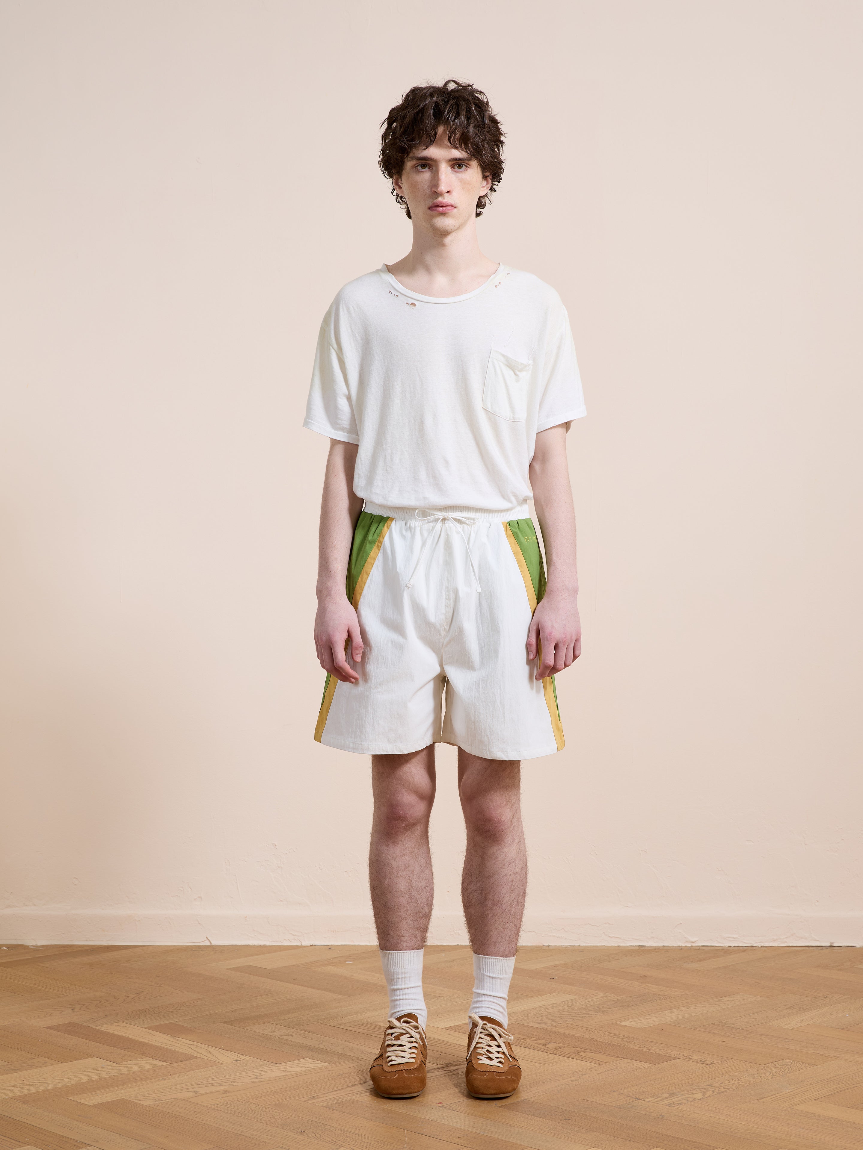 A person stands indoors against a plain wall, wearing a white t-shirt, white socks, brown shoes, and Found's Legacy Cricket Track Shorts with vintage-style green and yellow side panels.