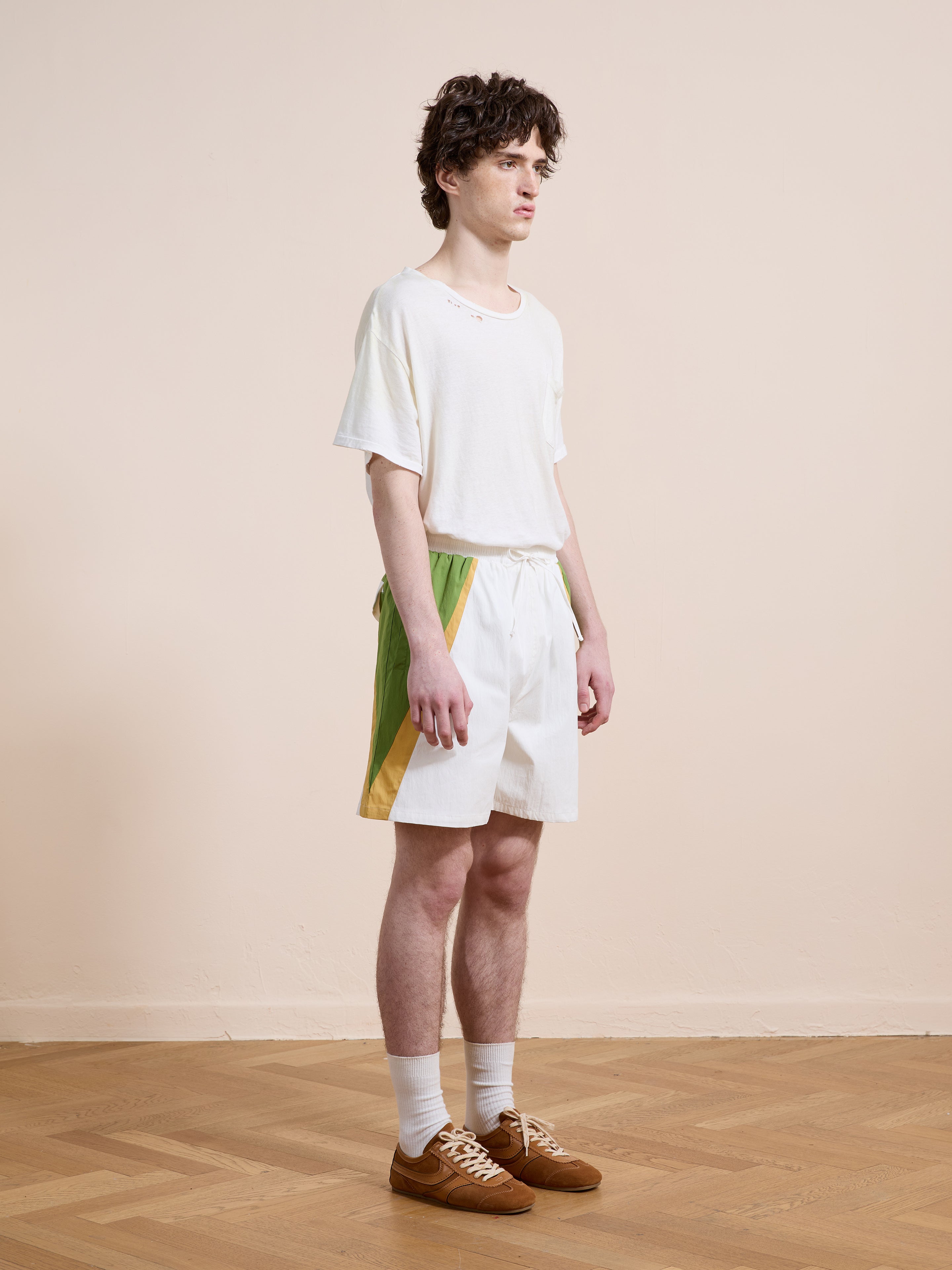 A young man stands indoors in a white tee and Found’s Legacy Cricket Track Shorts—white with green and yellow side panels—paired with white socks and brown sneakers, against a wooden floor and beige wall.