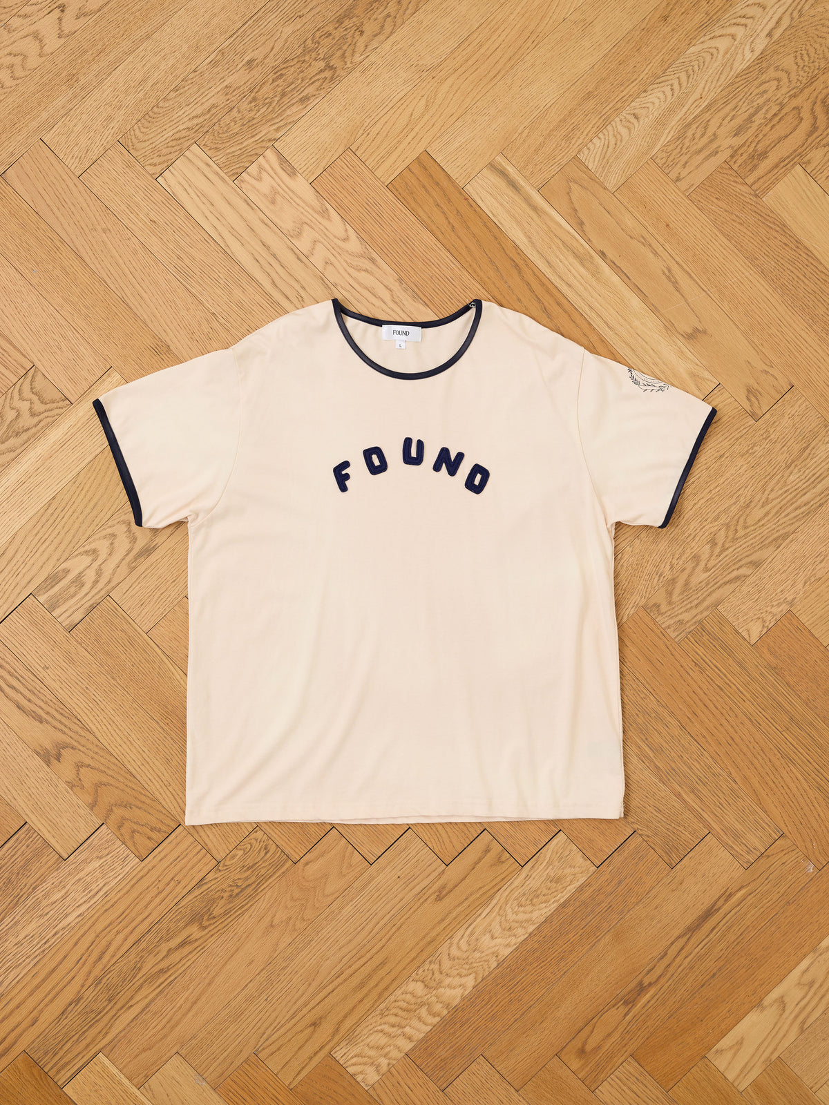 The Logo Felt Patch Ringer Tee by FOUND is a beige short-sleeve unisex style featuring black trim and a bold, embroidered “FOUND” logo across the chest. Displayed laid flat on a wooden herringbone floor, this relaxed fit tee combines comfort with timeless appeal.