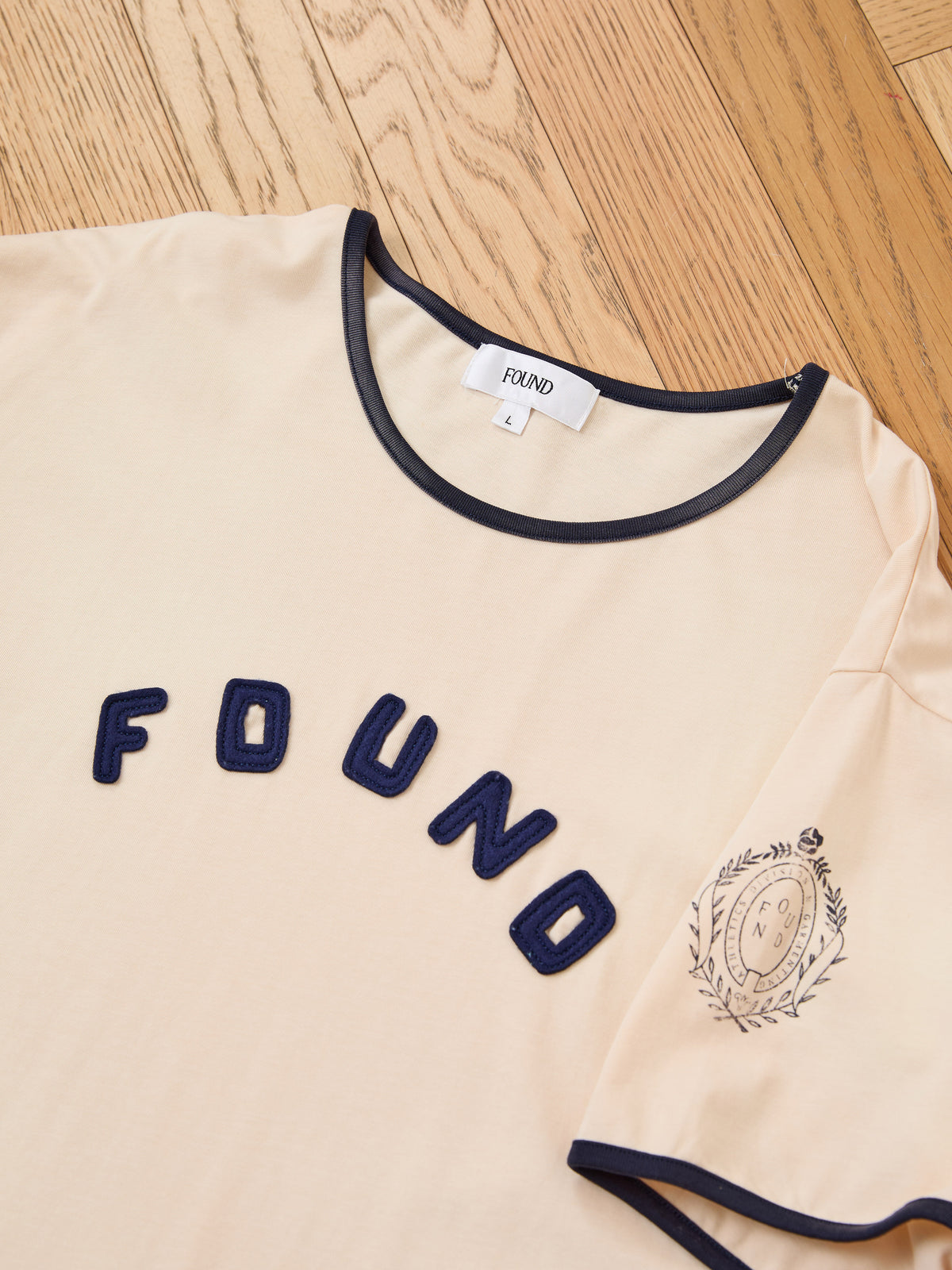 The FOUND Logo Felt Patch Ringer Tee in beige features a unisex relaxed fit with navy trim on the collar and sleeves, “FOUND” lettering in navy felt across the chest, a small crest detail on the left sleeve, and is displayed laid flat on a wooden floor.