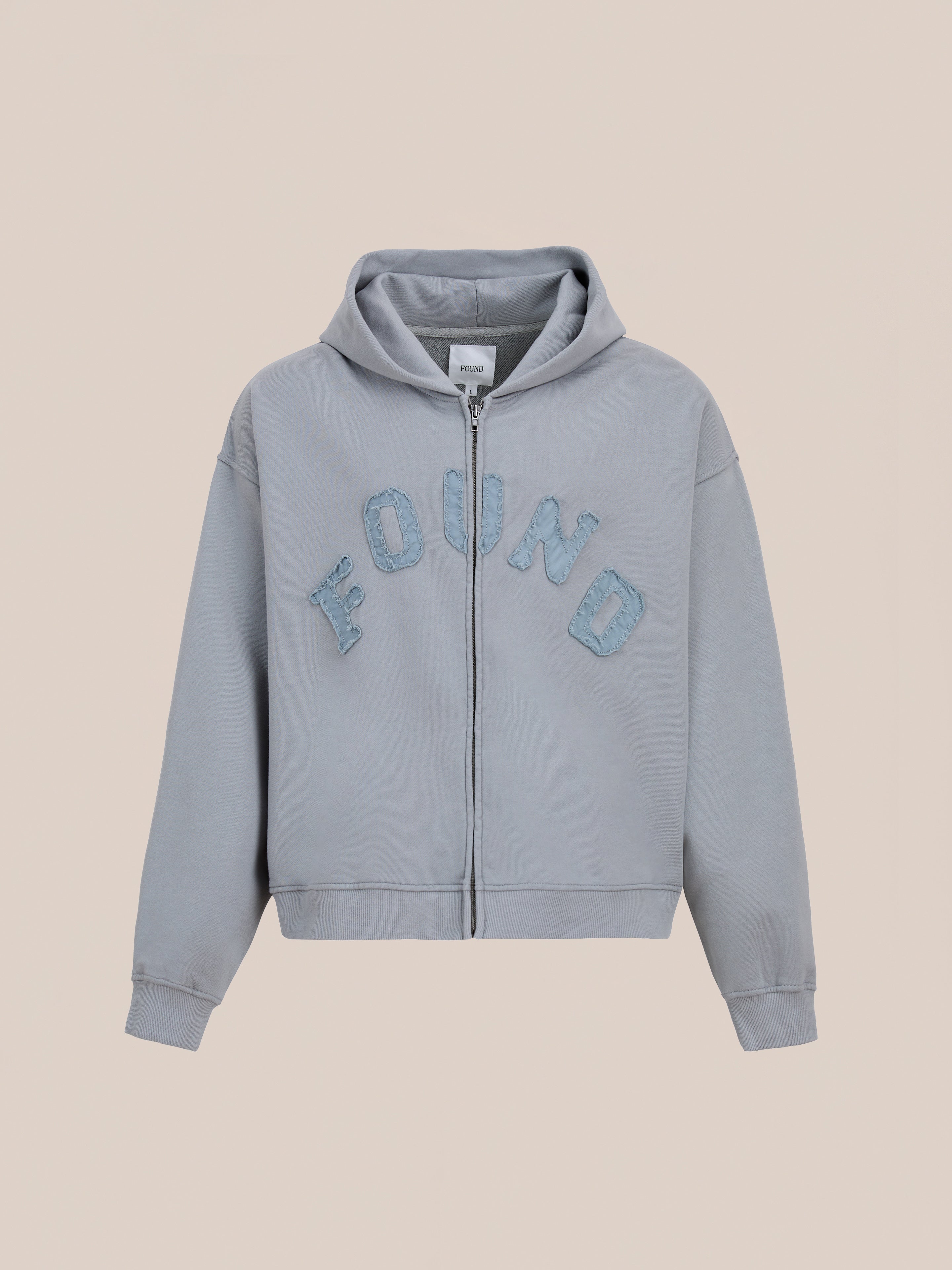 The FOUND Logo Patch Hoodie is a washed gray, unisex zip-up with a hood and bold textured blue "FOUND" lettering on the chest; relaxed fit ideal for vintage athletic style, shown on a plain beige background.