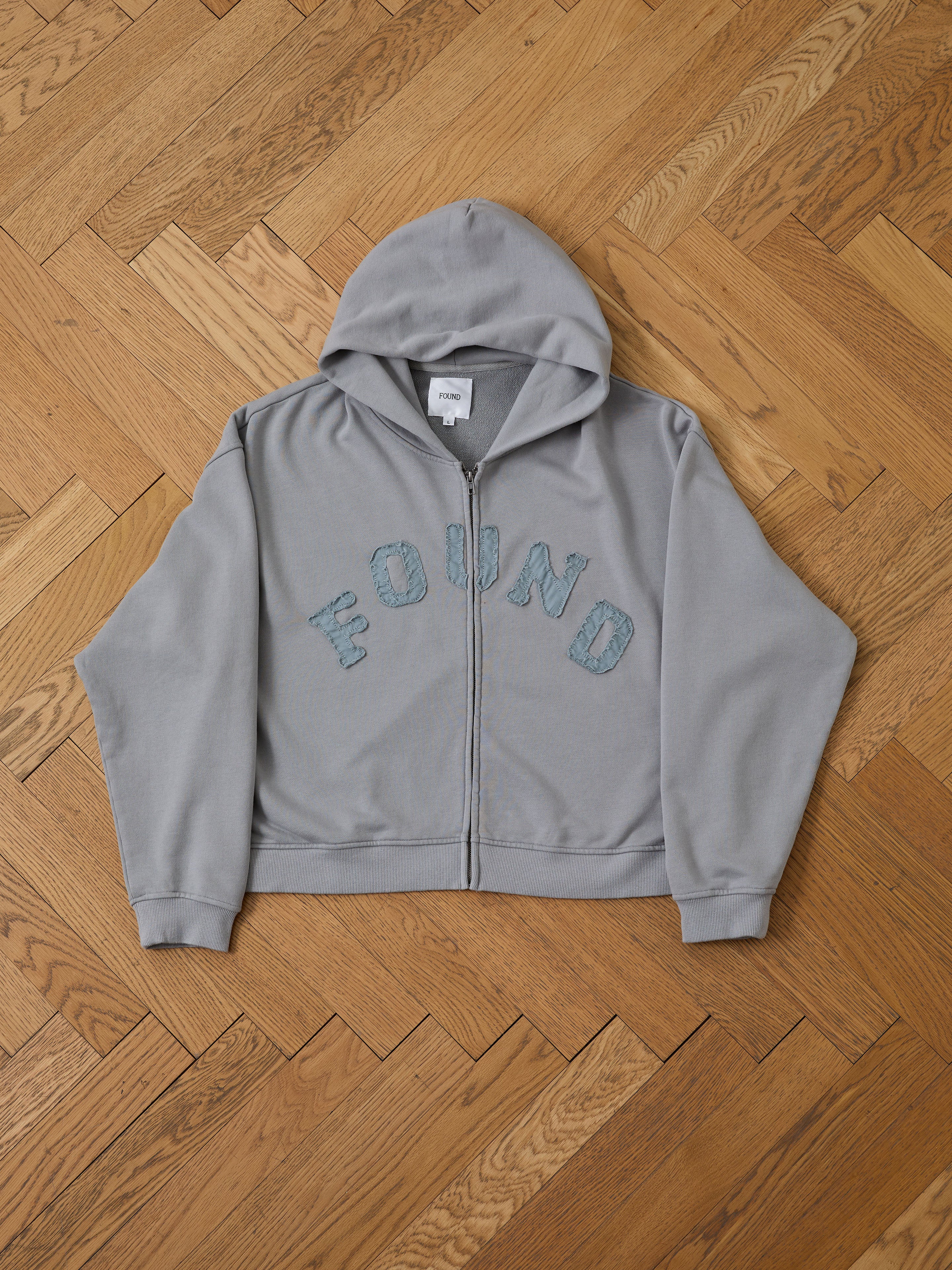 The FOUND Logo Patch Hoodie is a washed gray zip-up with a hood, shown flat on wood. "FOUND" appears in bold blue letters on the chest—ideal for unisex style and vintage athletic layering.