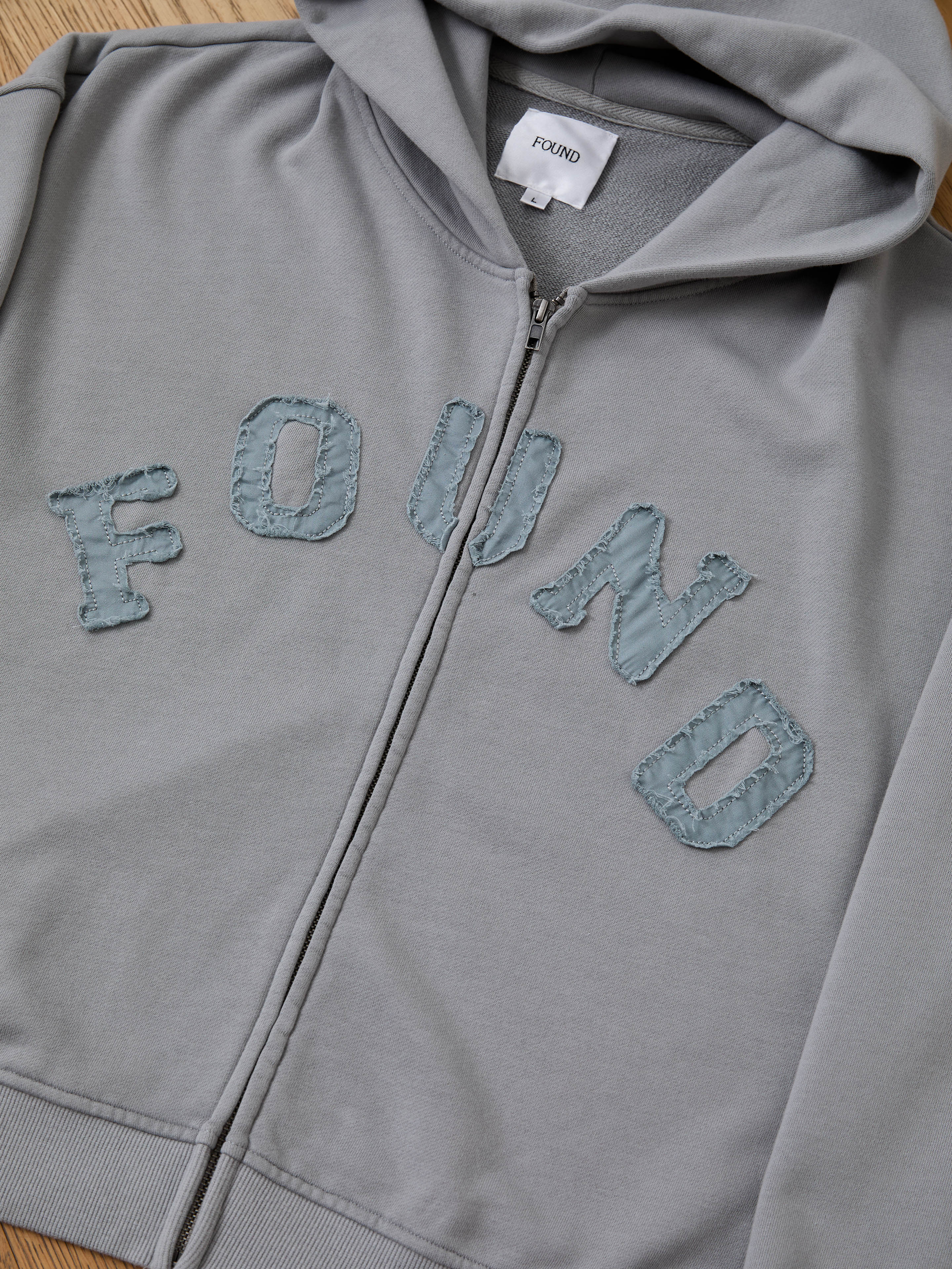 The FOUND Logo Patch Hoodie is a gray unisex relaxed fit zip-up featuring a hood and large textured blue "FOUND" letters sewn on the front for a vintage athletic style.