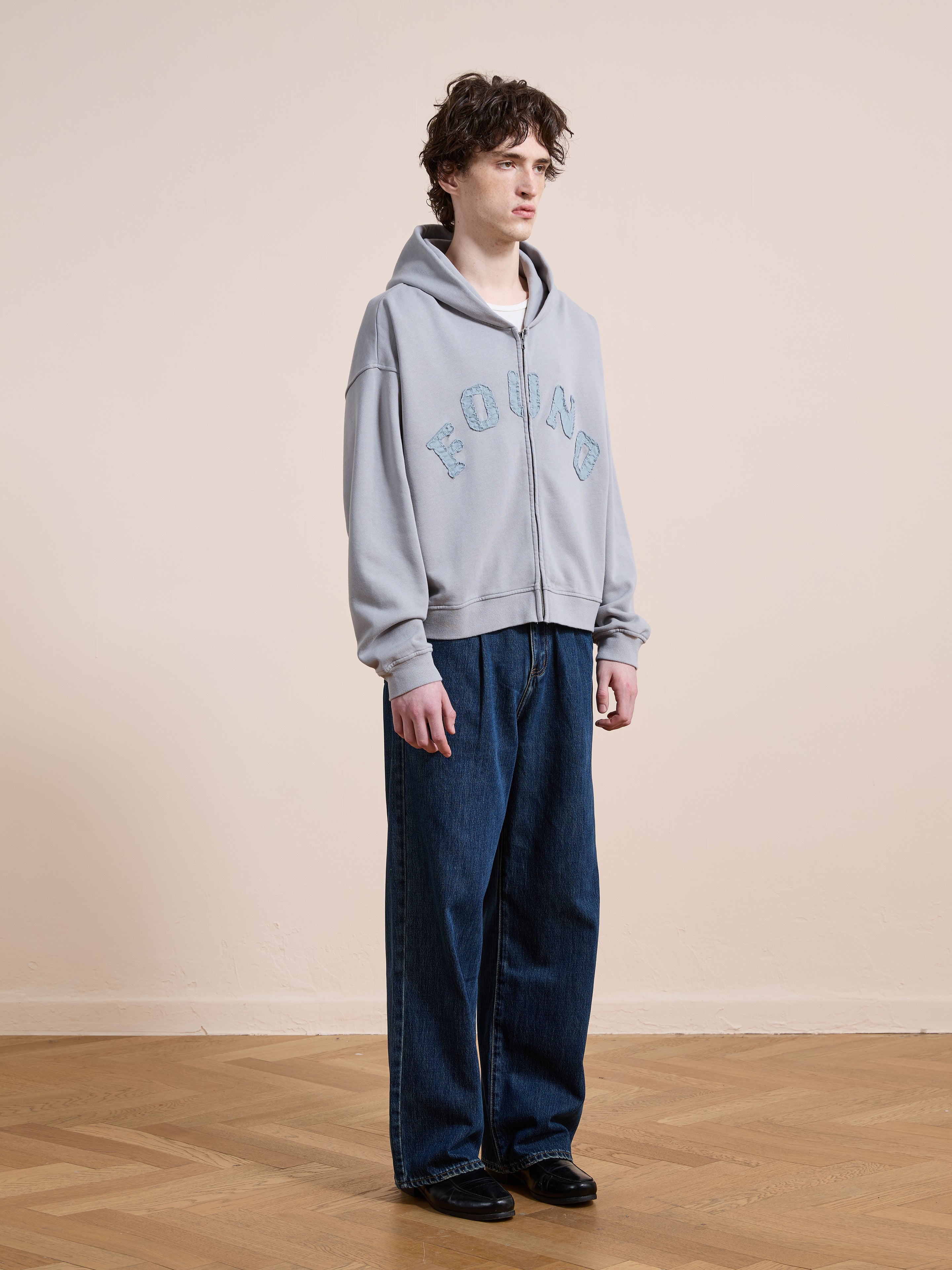 A person stands indoors on a wooden floor in a FOUND Logo Patch Hoodie, unisex relaxed fit blue jeans, and black shoes, against a plain beige wall.