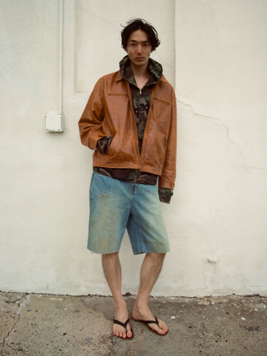 A man stands against a light-colored wall wearing the FOUND Marlboro Sun Faded Leather Racer Jacket, a camouflage hoodie, denim shorts, and black flip-flops.
