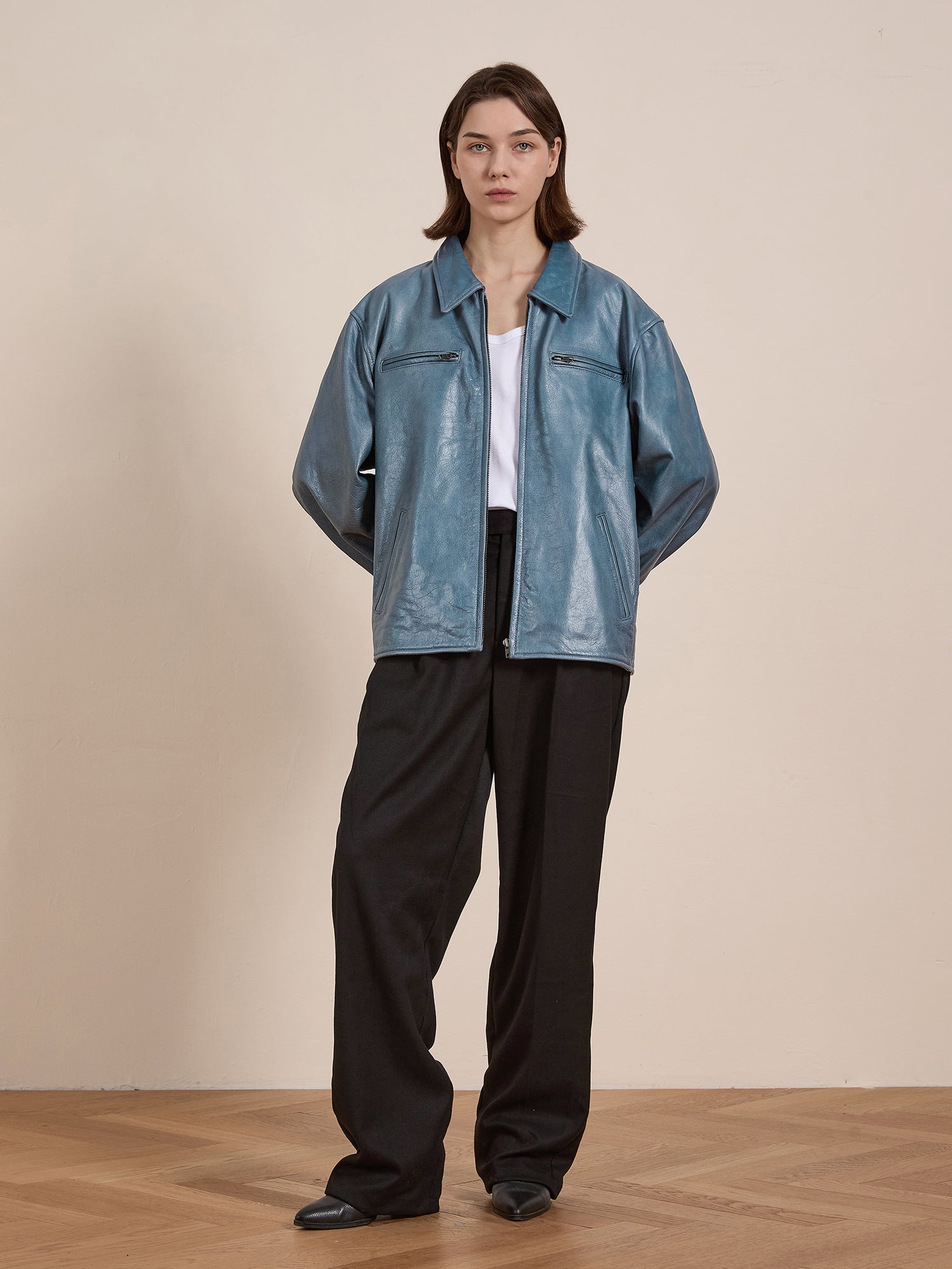 A person wears the Marlboro Sun Faded Leather Racer Jacket by FOUND, featuring a light blue unisex relaxed fit. They pair it with a white shirt and black pants while standing on a wooden floor against a plain background.