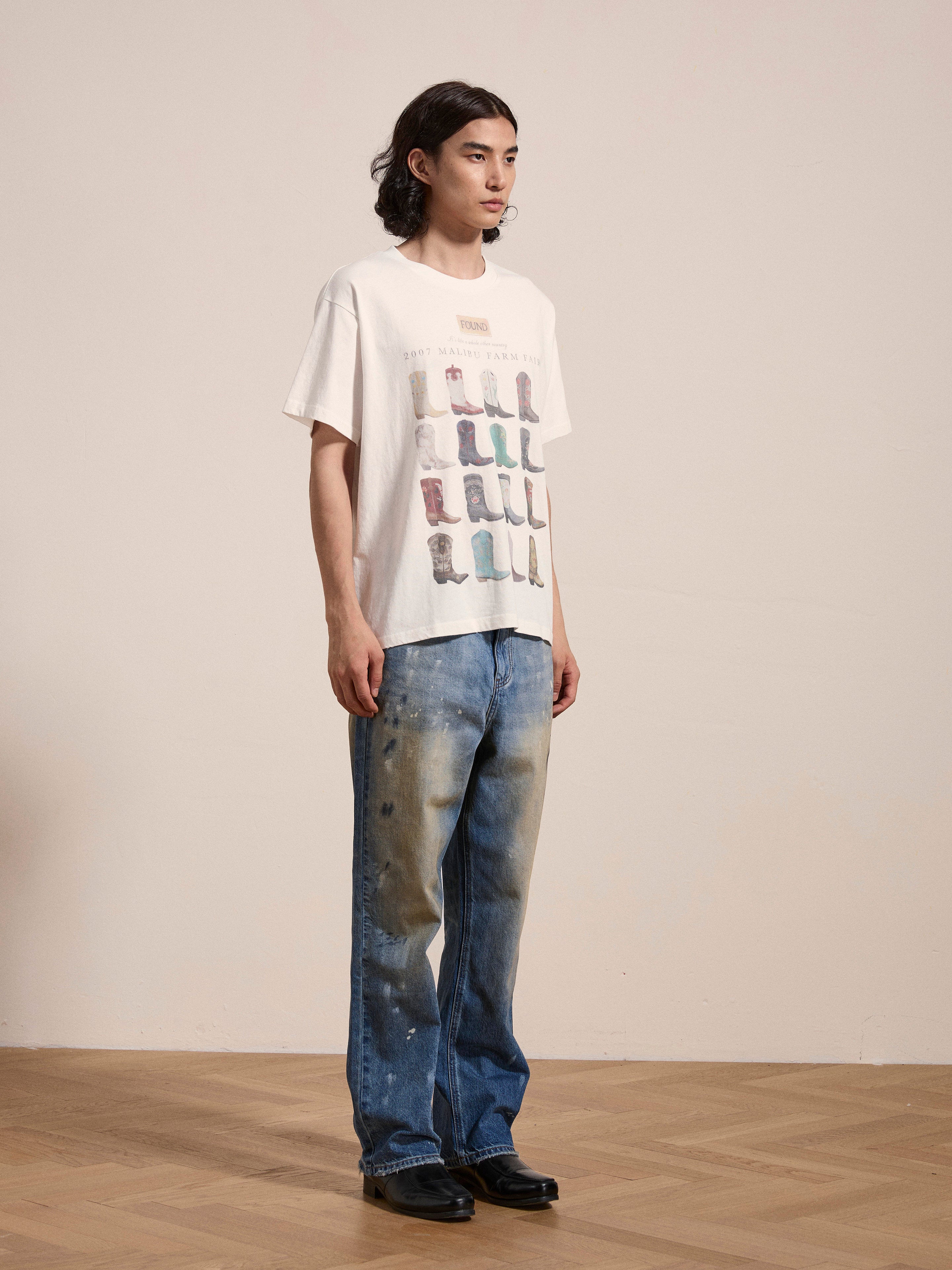 A person with long hair wears a white Malibu Farm Fair Tee from FOUND, made from 100% cotton, and distressed blue jeans, standing on a hardwood floor against a plain background.