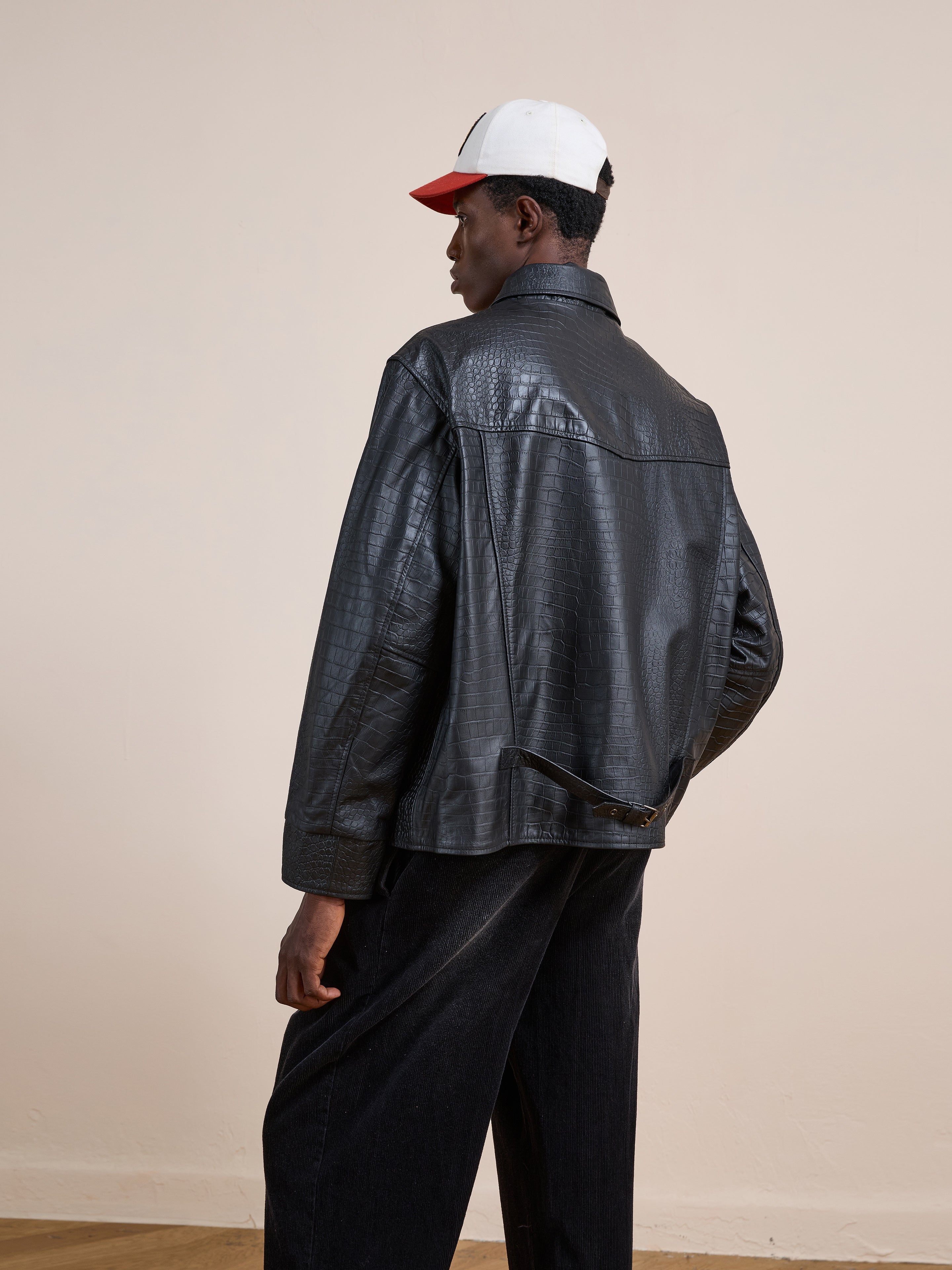 A person faces away, wearing the FOUND Marlboro Croc Embossed Leather Racer Jacket in a unisex relaxed fit, paired with dark pants and a white cap with a red brim, standing against a plain light background.