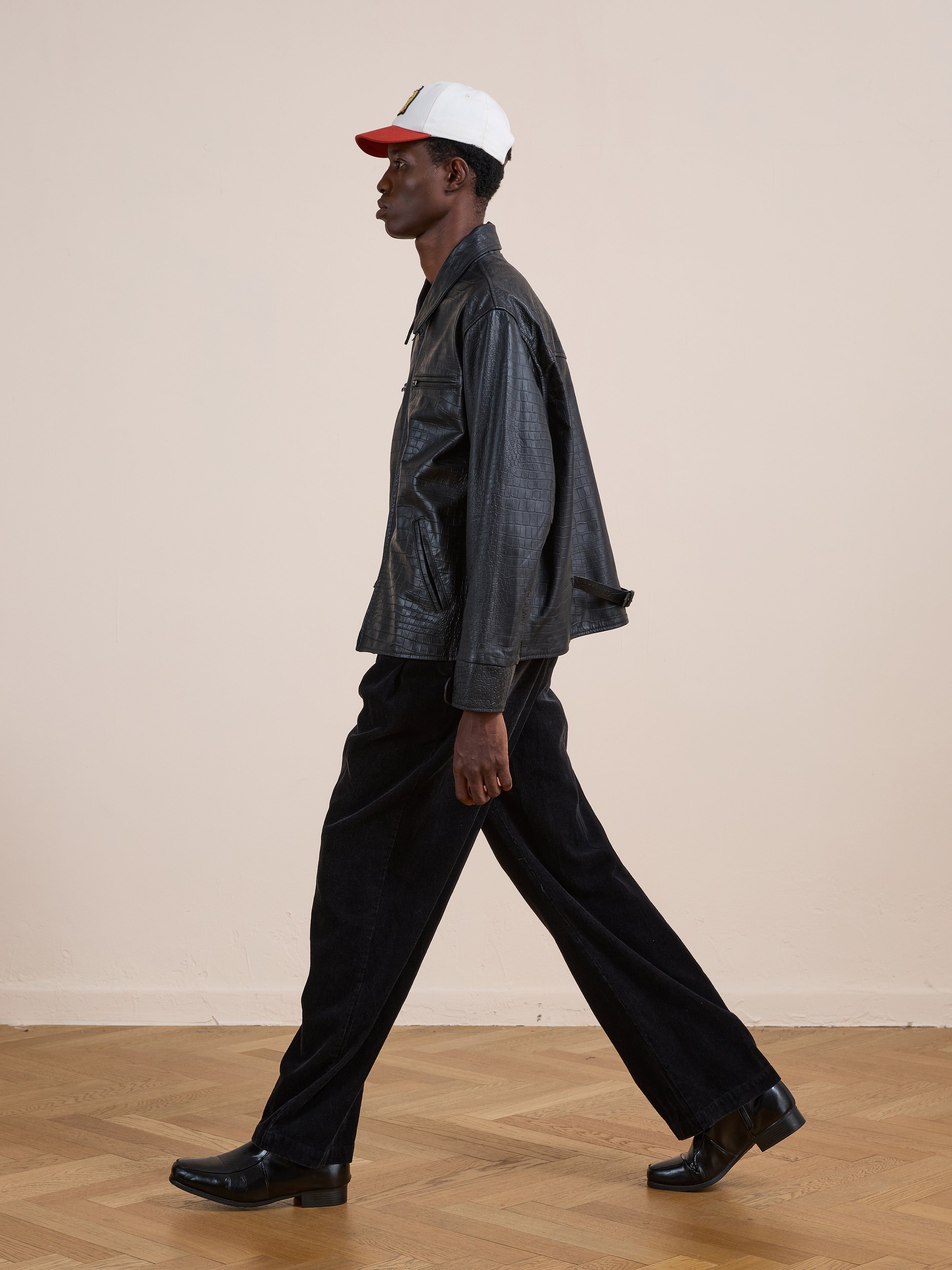 A person wearing the FOUND Marlboro Croc Embossed Leather Racer Jacket, black pants, black shoes, and a white and red cap walks indoors on a wooden floor.