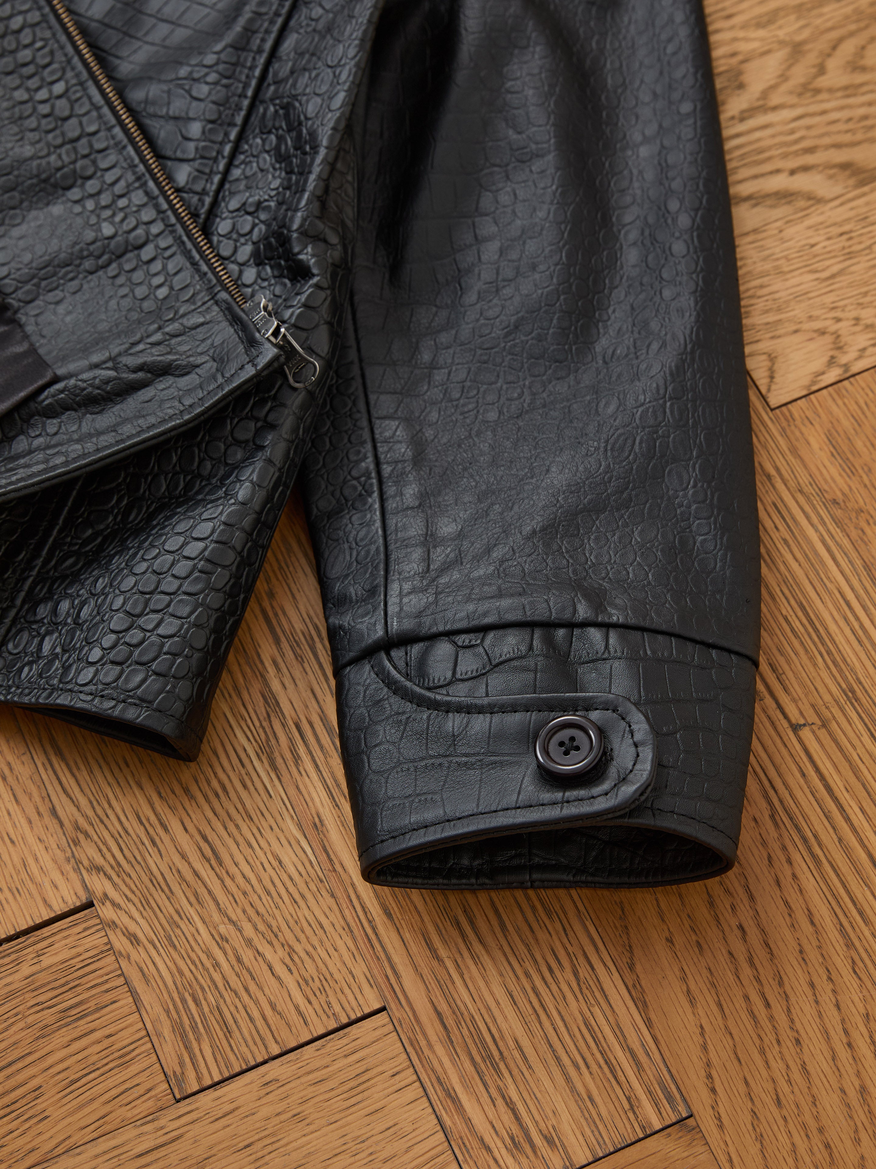 Close-up of the FOUND Marlboro Croc Embossed Leather Racer Jacket sleeve, featuring black faux snakeskin, a buttoned cuff, and visible zipper in a unisex relaxed fit, displayed on a wooden floor.