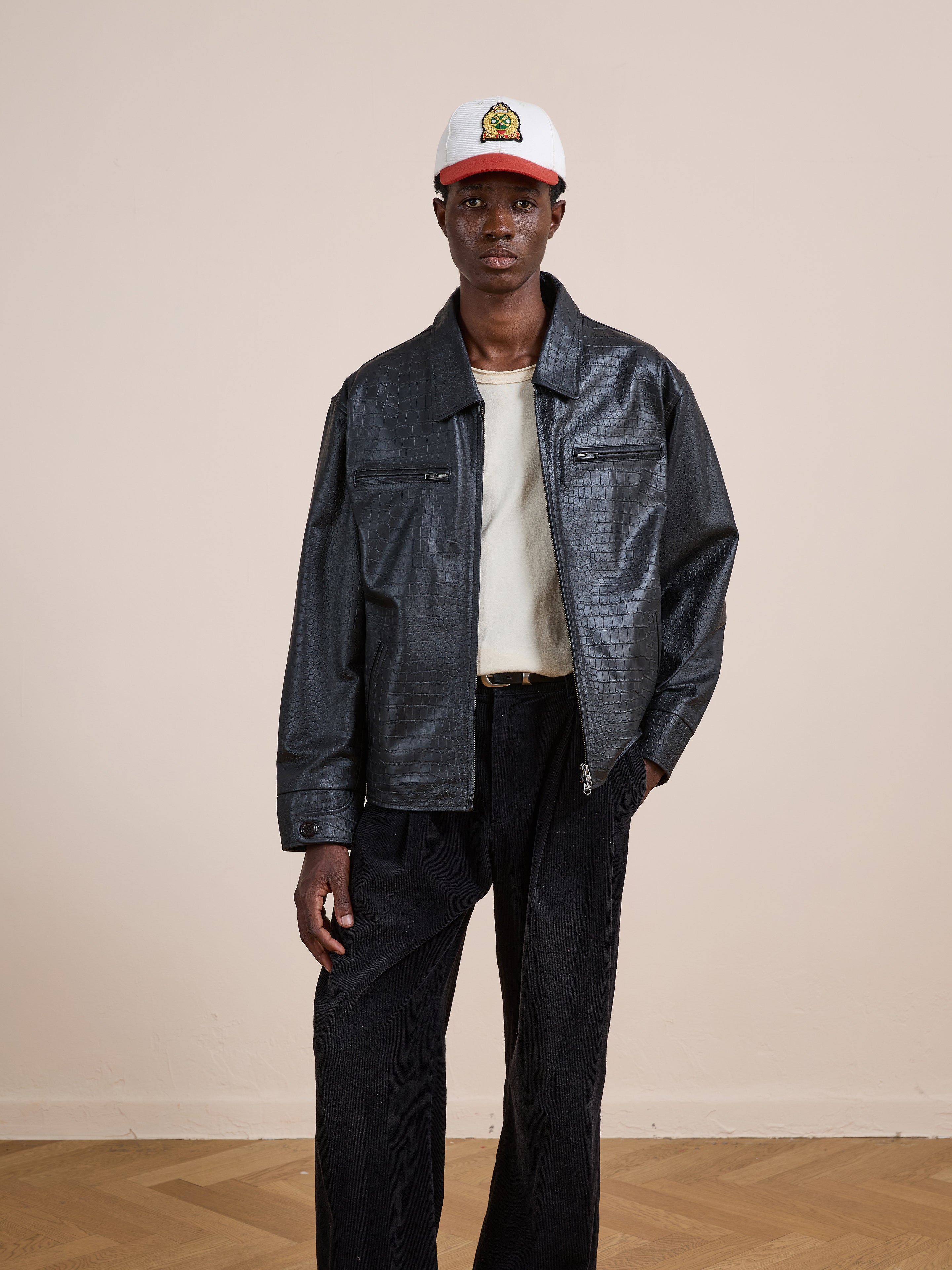 A person stands indoors wearing the FOUND Marlboro Croc Embossed Leather Racer Jacket, a white cap, off-white t-shirt, and black pants, posing neutrally against a beige wall.