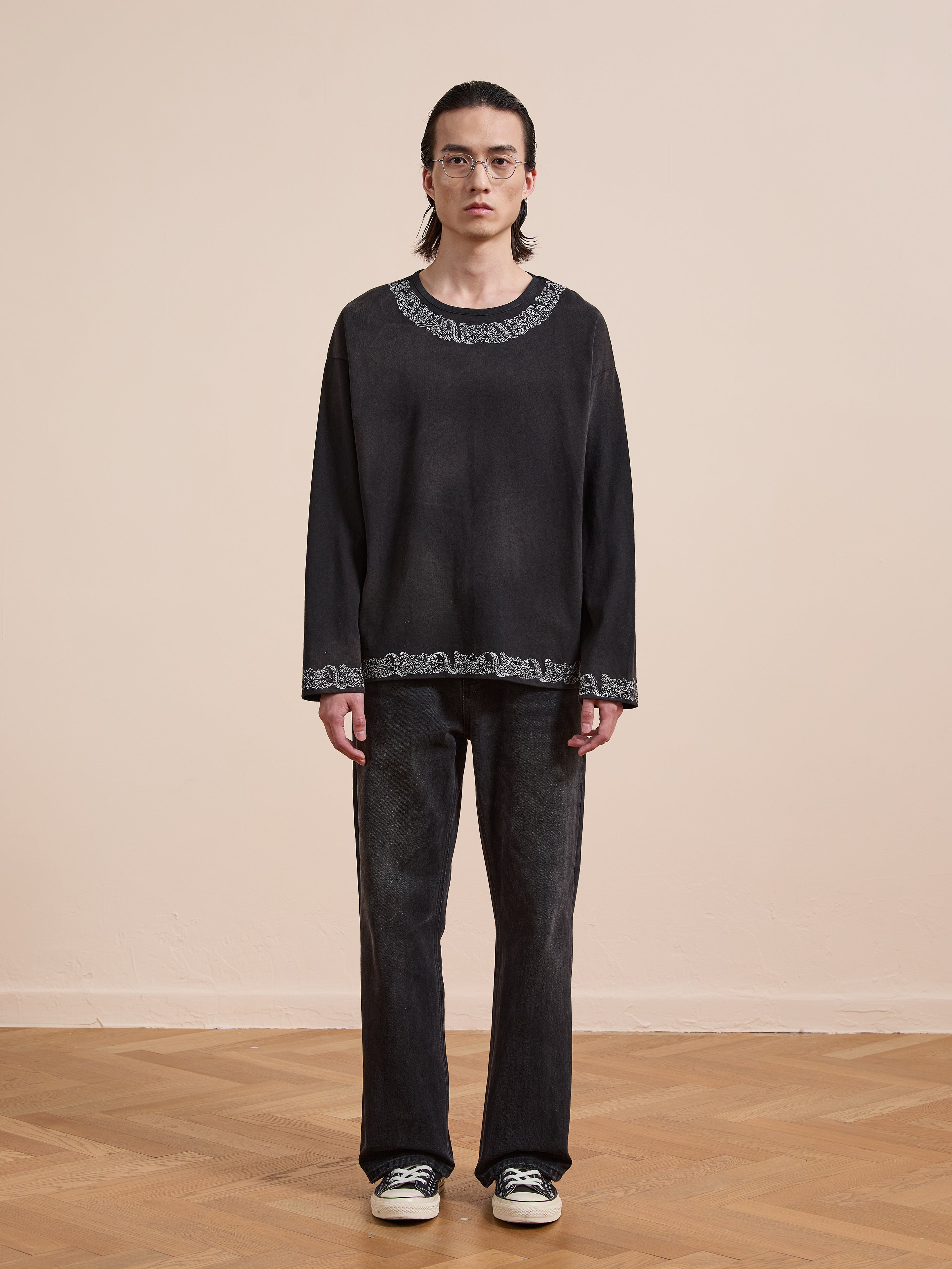 A person stands indoors on a wooden floor wearing glasses, dark jeans, black-and-white sneakers, and the FOUND Metallic Long Sleeve Embroidered Tee—a vintage-style unisex relaxed fit shirt with decorative trim.