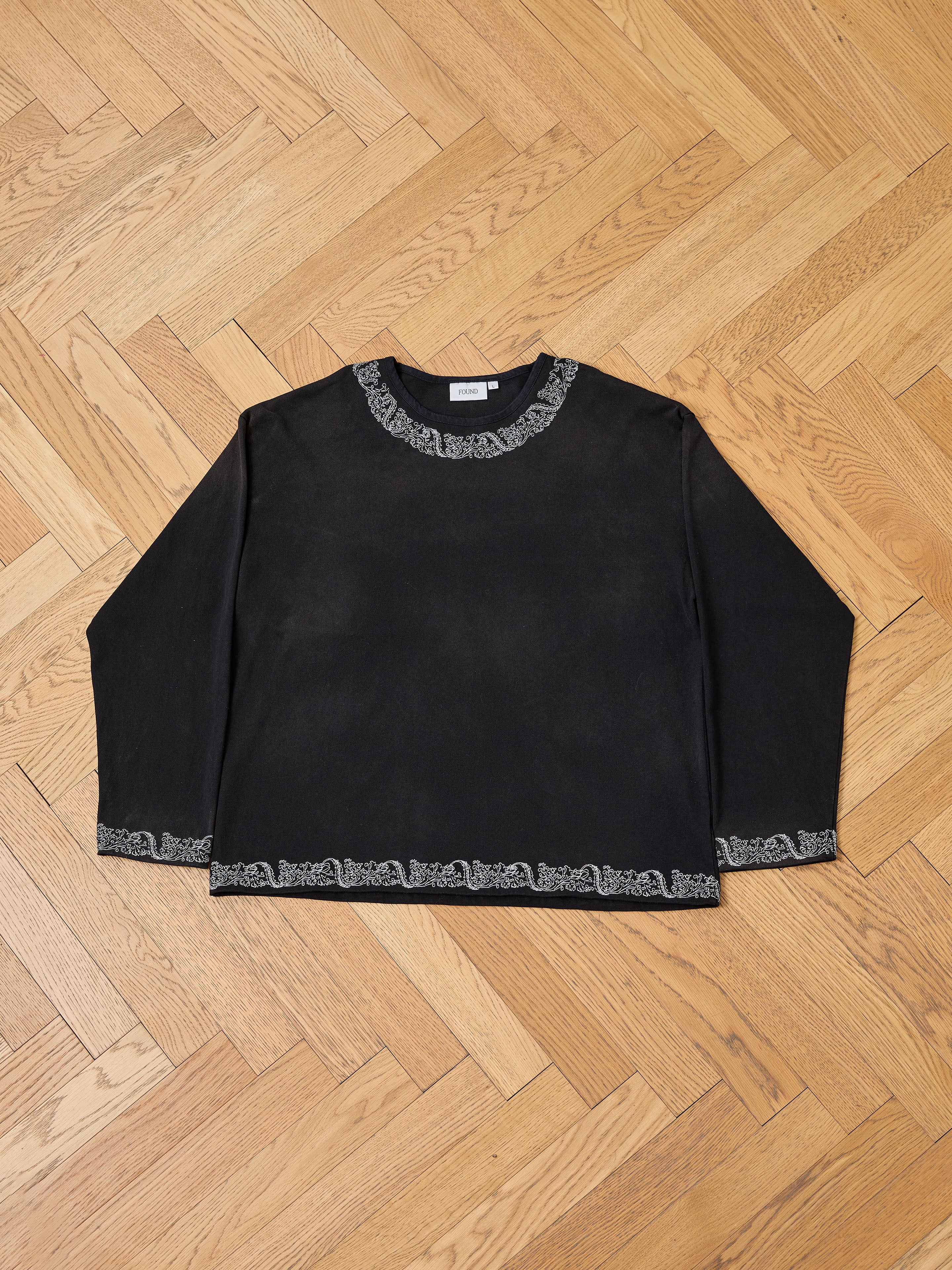 The FOUND Metallic Long Sleeve Embroidered Tee, a vintage black unisex relaxed fit shirt with gray patterned trim on the collar, cuffs, and hem, is shown laid flat on a wooden parquet floor.