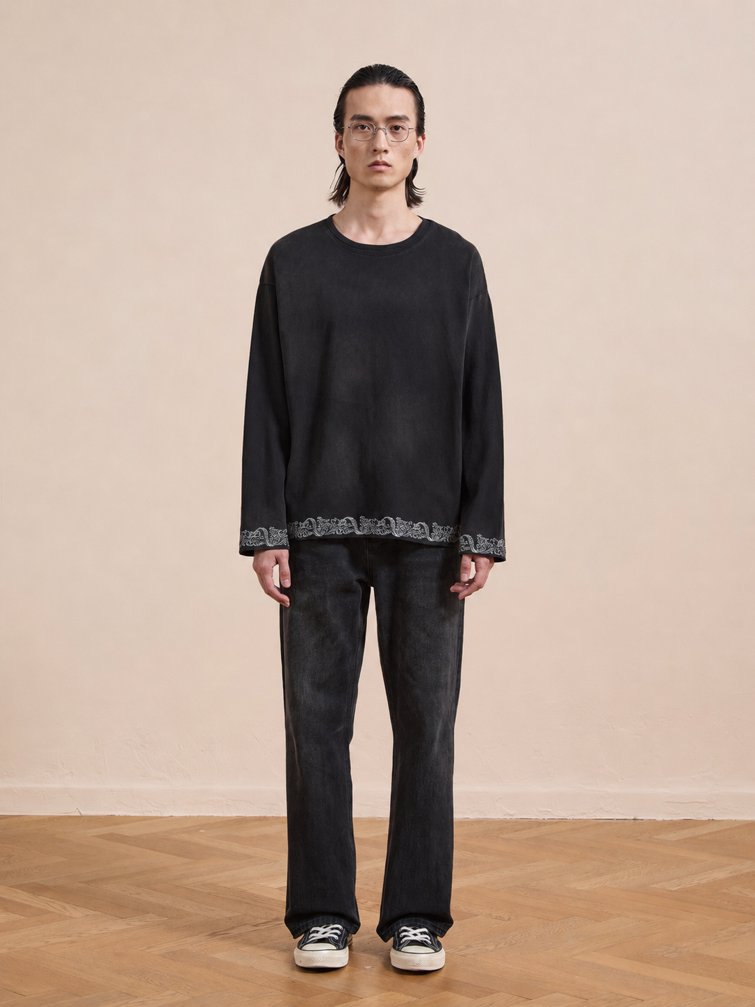 A person stands indoors on a wooden floor, facing the camera against a beige wall, wearing the FOUND Metallic Long Sleeve Embroidered Tee with black pants and black-and-white sneakers.