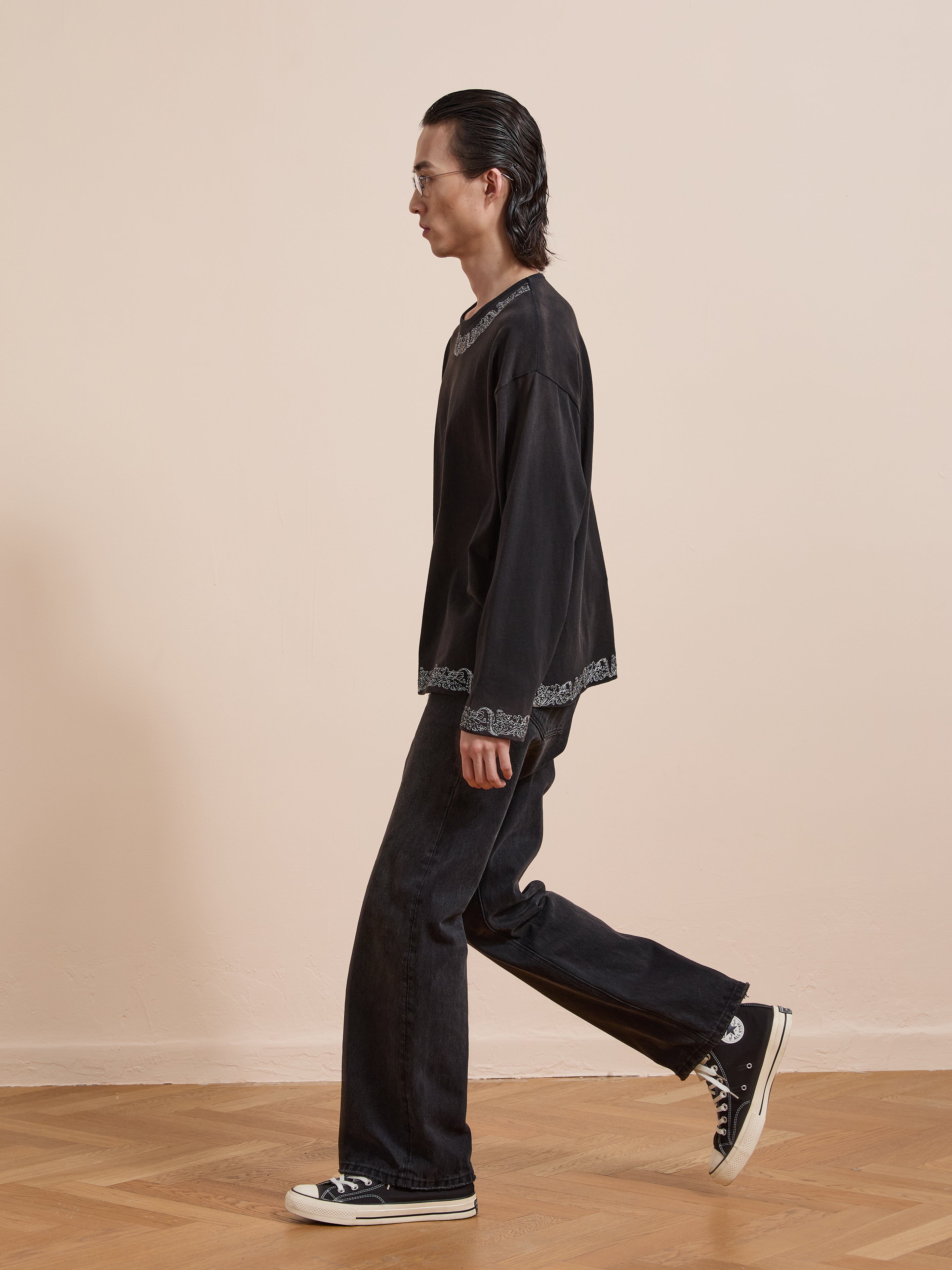 A person with long dark hair and glasses, dressed in a FOUND Metallic Long Sleeve Embroidered Tee, black jeans, and black Converse sneakers, walks indoors on a wooden floor against a beige wall.